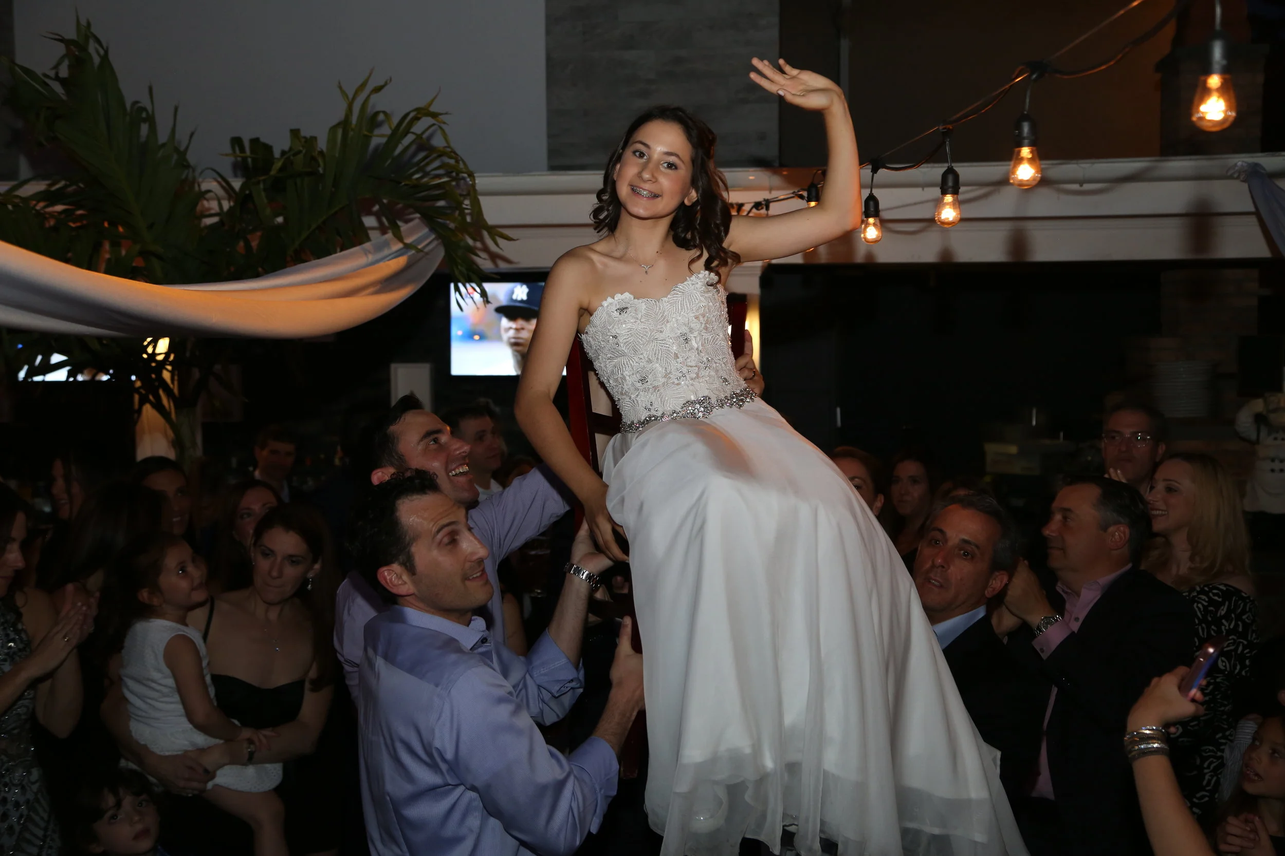 A young woman in a white wedding dress is being lifted above a crowd of people at a celebration or wedding reception, smiling and waving.