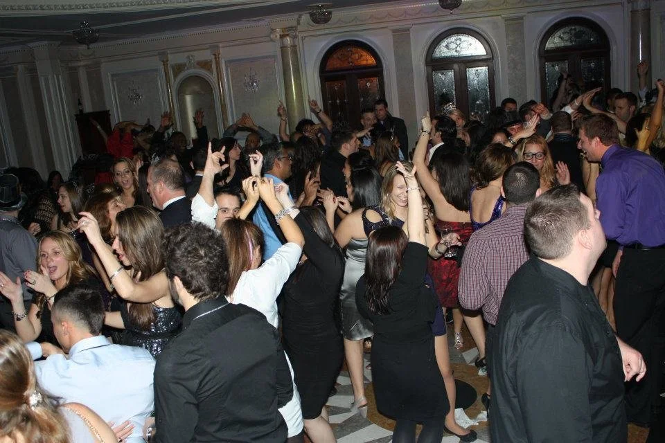 Crowd of people dancing and celebrating at a party in a decorated ballroom with ornate walls and arched windows.