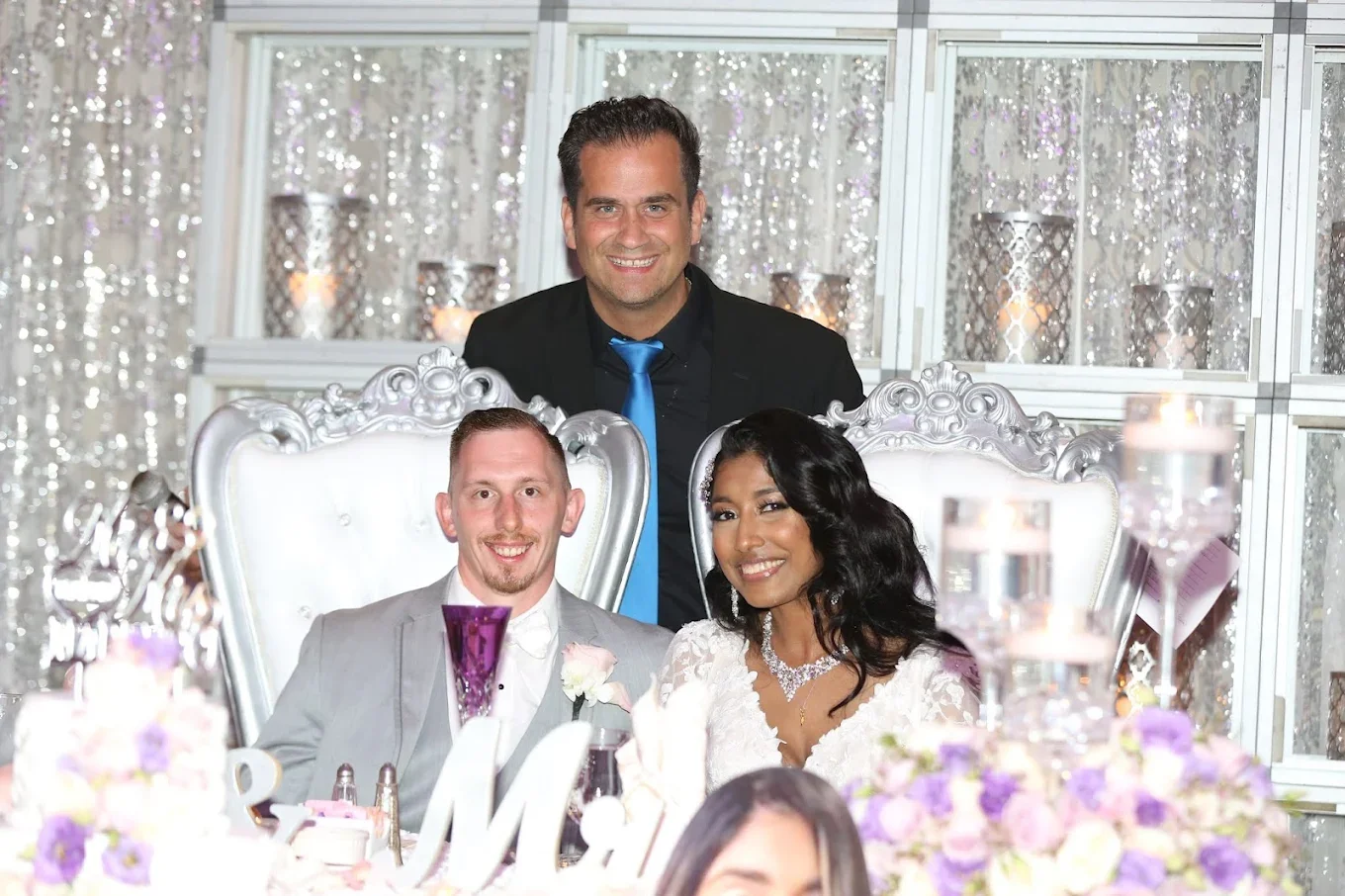 A man and woman sitting at a wedding reception table with another man standing behind them. The woman is wearing a white wedding dress and the man is in a light gray suit. The man standing is wearing a black suit and a blue tie. The table is decorate