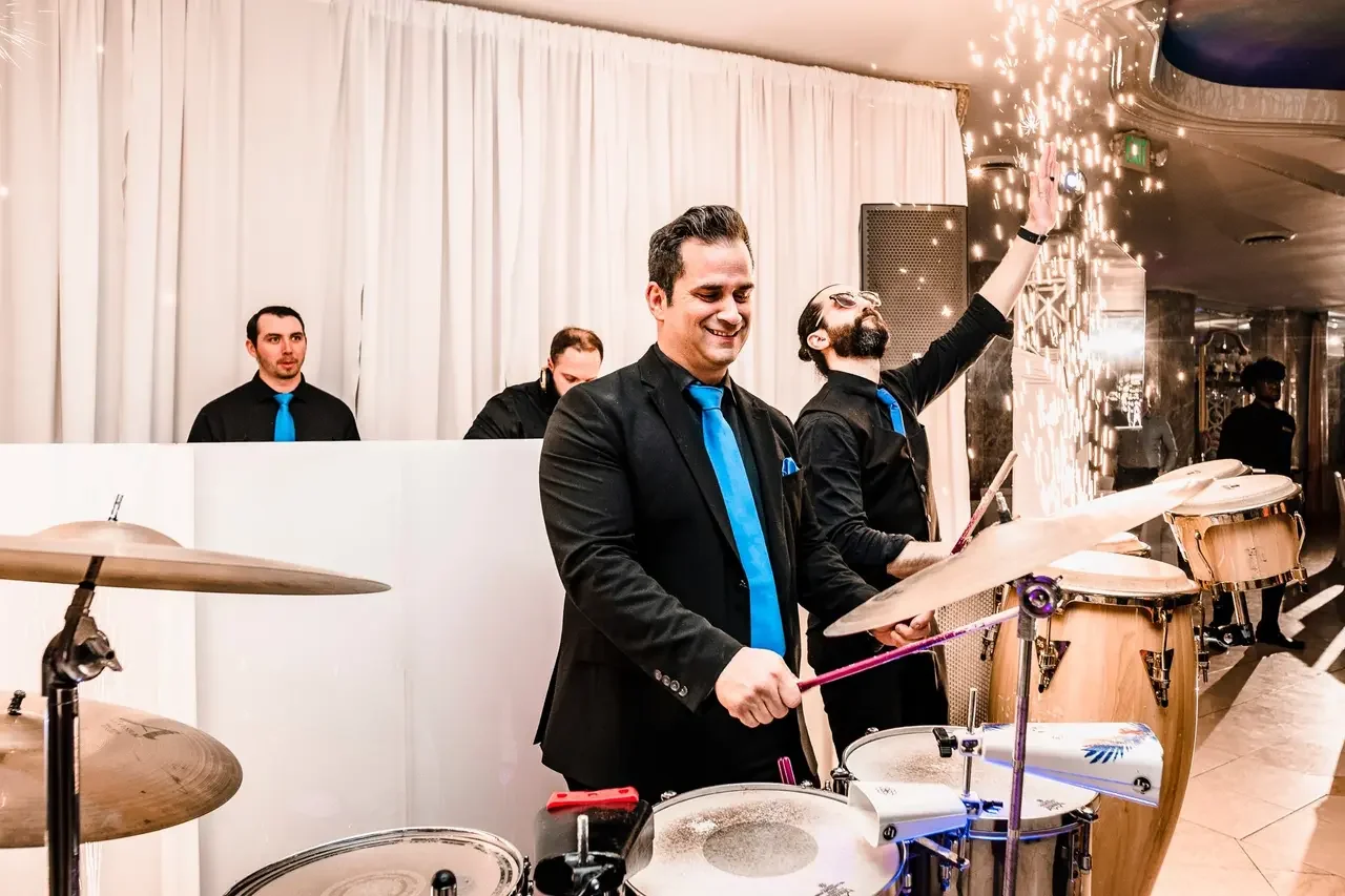Group of musicians playing percussion instruments at an indoor event with white drapery and celebratory lighting.