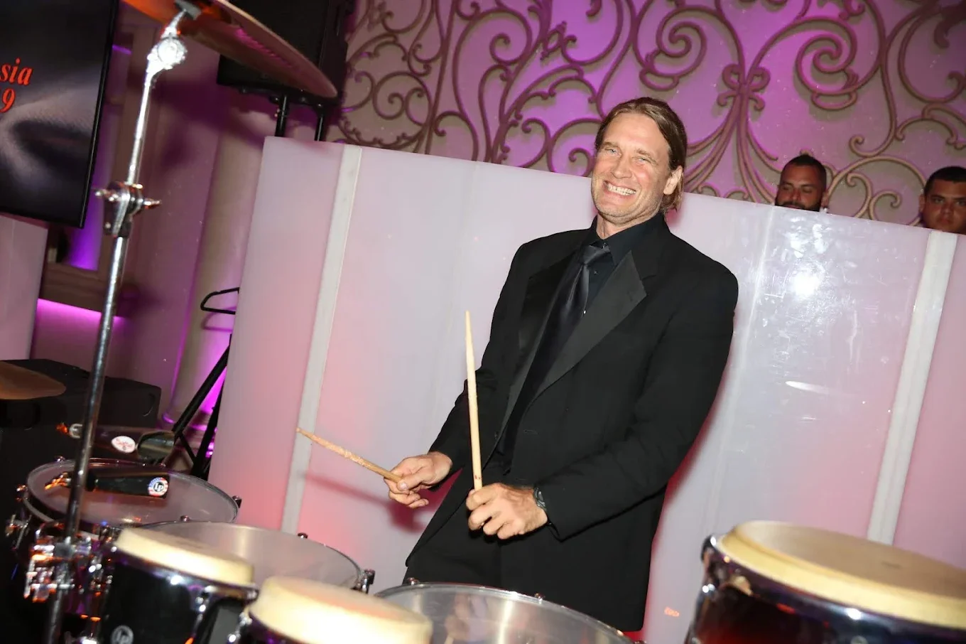 A man in a black tuxedo smiling and playing drums at a formal event with pink and purple background decor.