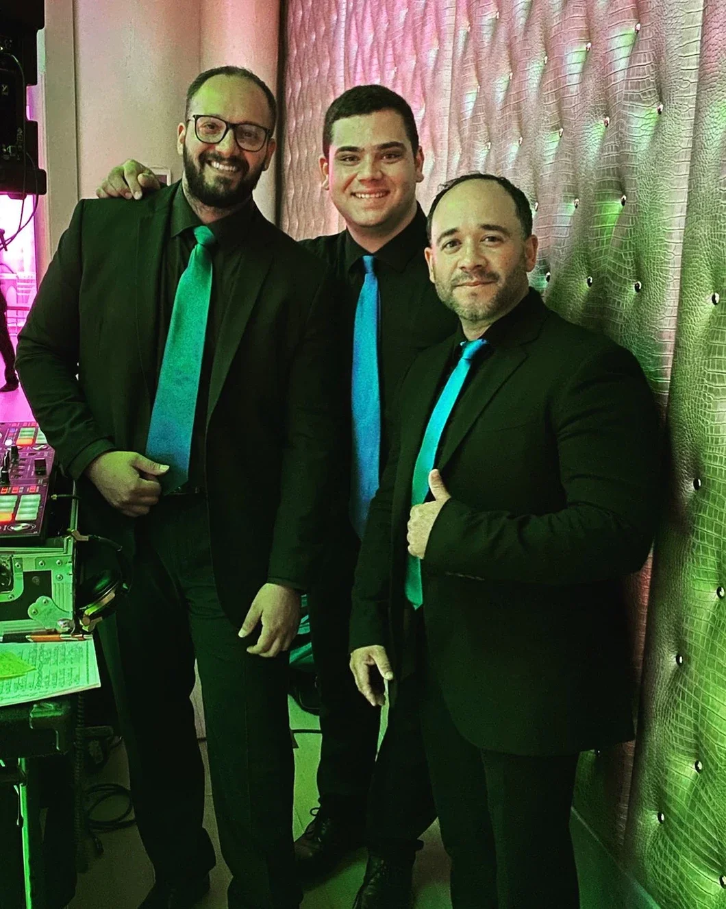 Three men dressed in black suits and colorful ties, standing indoors against a patterned wall with pink and green lighting, smiling at the camera.