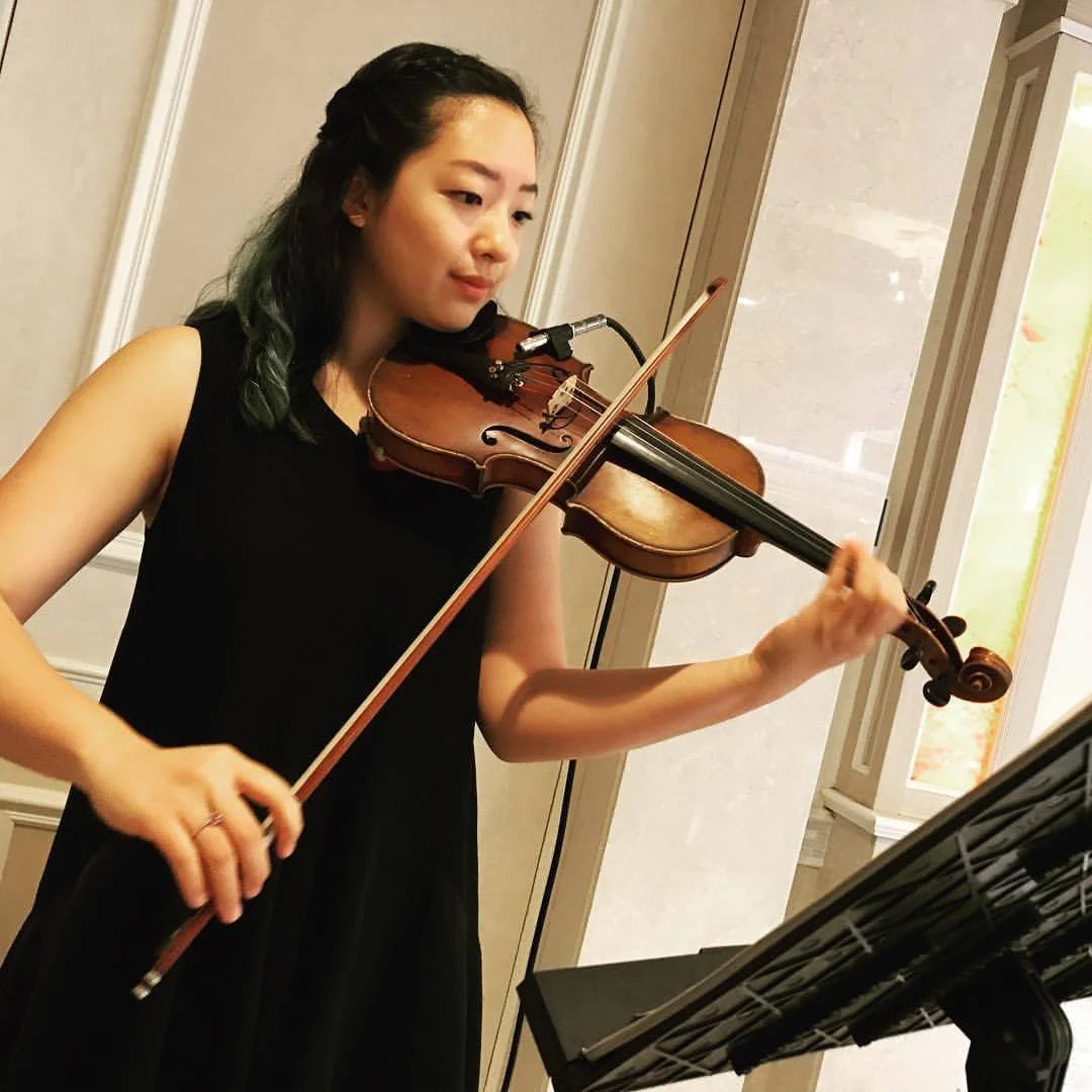 A young woman with dark hair and green streaks, wearing a black sleeveless dress, playing a violin while reading sheet music on a stand in a room with beige walls and a window.
