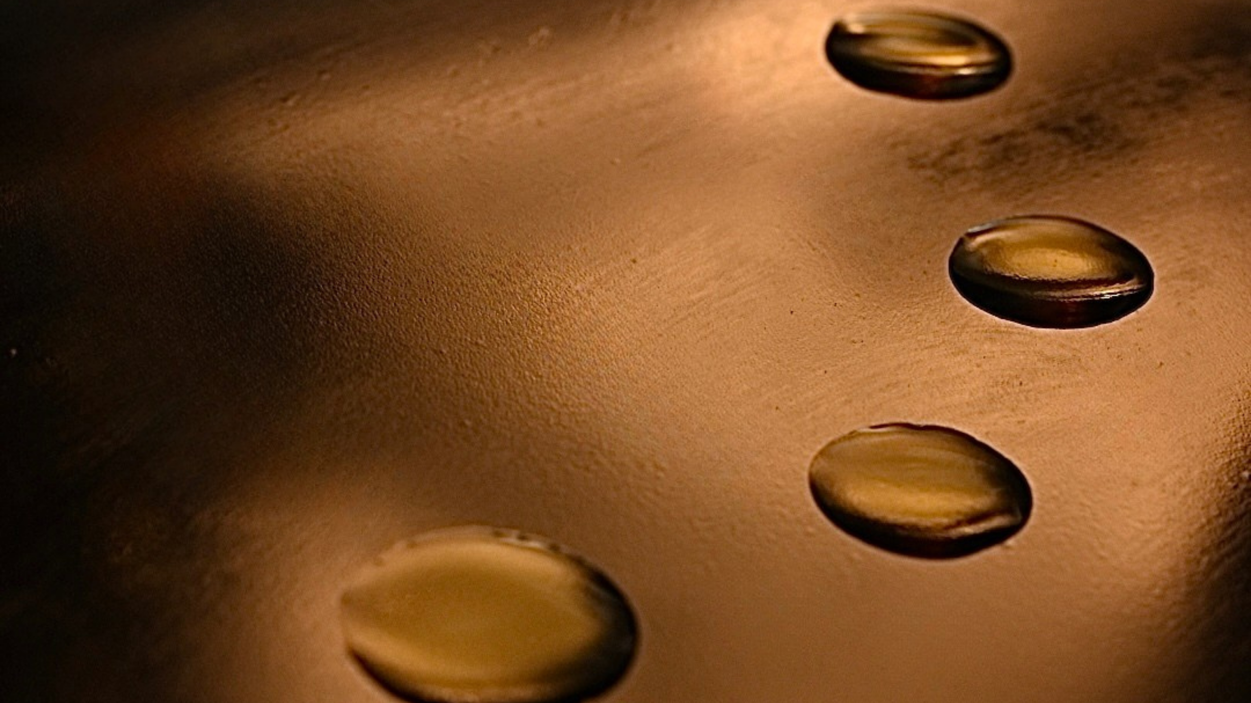 Close-up of Honeybeam, Golden Treatment nectar, which ia an oil on a smooth, metallic surface illuminated with warm lighting