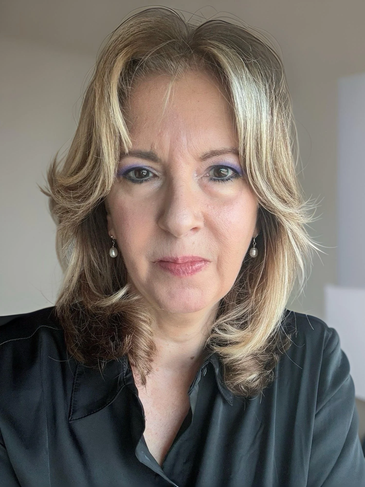 A woman with blonde, wavy hair wearing pearl earrings and a black top, taking a selfie indoors with a neutral wall background.