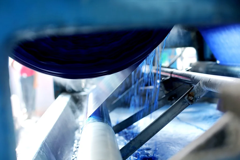 Close-up of a manufacturing process involving a large black belt or roll, with blue liquid or material flowing over a metal surface, in an industrial setting.