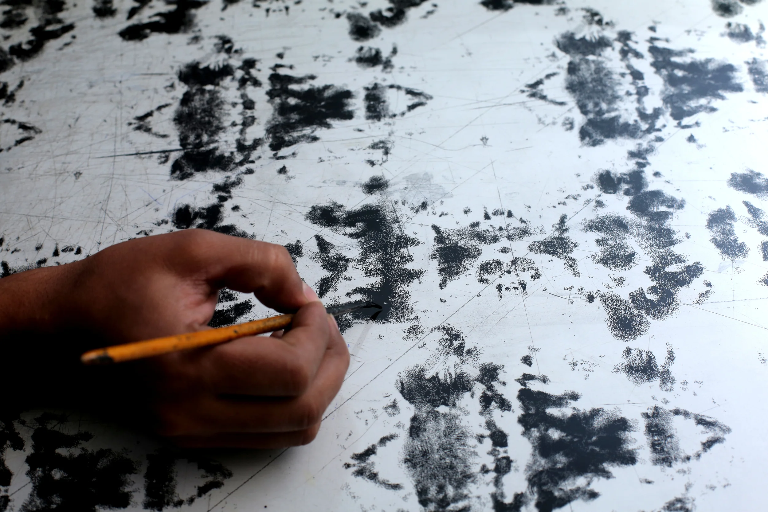 Close-up of a hand holding a paintbrush, creating black ink or pigment marks on a white surface, with various textured black patterns and lines.