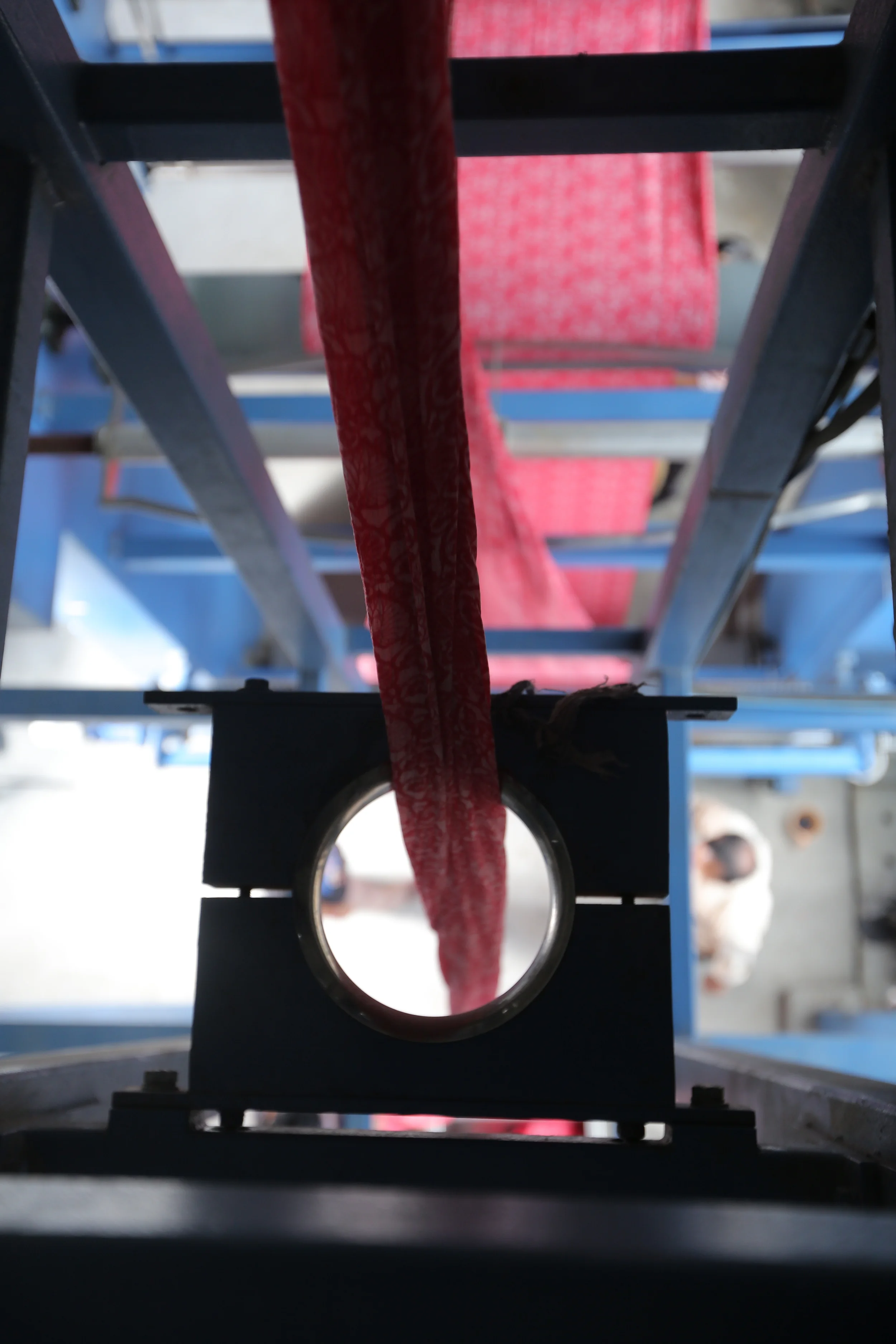 Close-up of a mechanical part with a red and white fabric passing through a metal ring, part of a testing or manufacturing setup, with a blurred dog in the background.