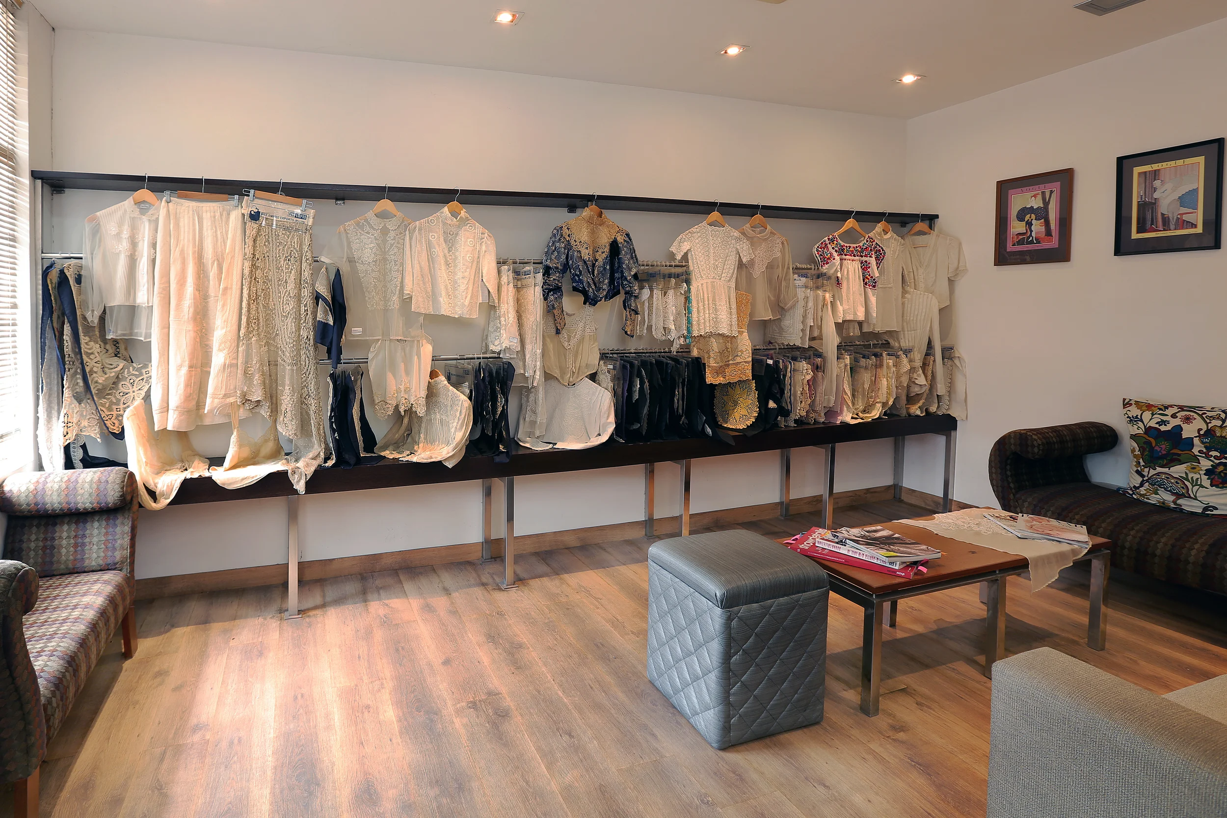 Clothing displayed on a rack with dresses, blouses, and skirts, inside a room with wooden flooring, couches, a coffee table with magazines, and framed artwork on the wall.