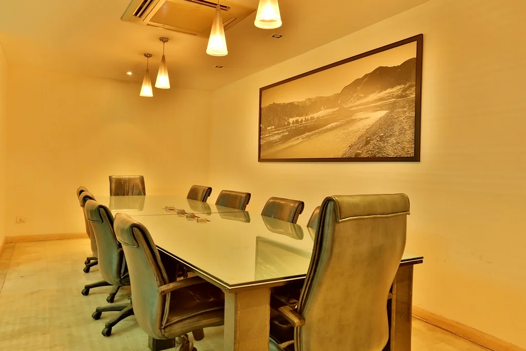 Empty conference room with a large glass table and eight green leather chairs, a black-and-white landscape photograph on the wall, and three hanging pendant lights.