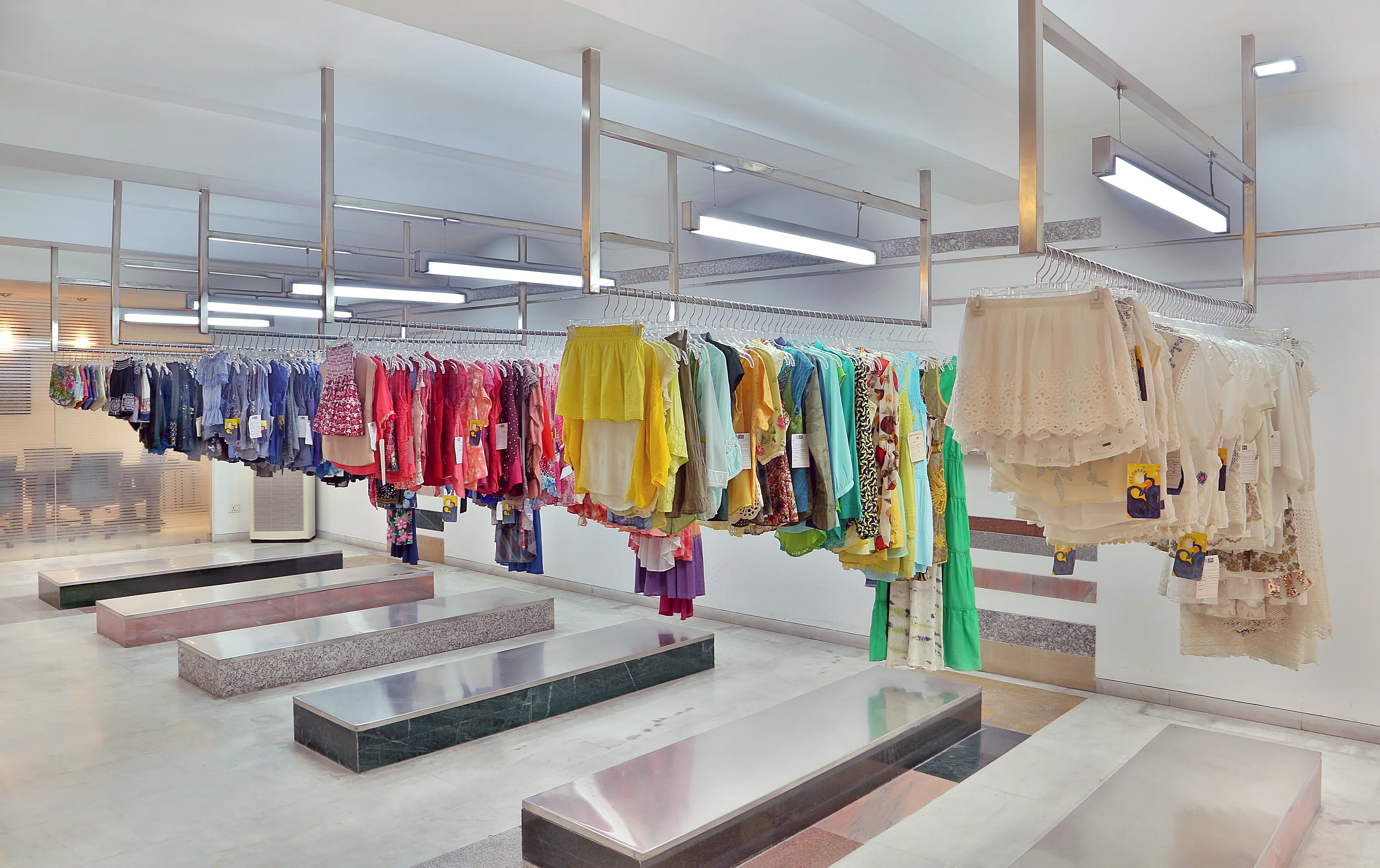 Clothing store with colorful women's clothes hanging on metal racks, displays of dresses and skirts, and store benches on a marble floor.