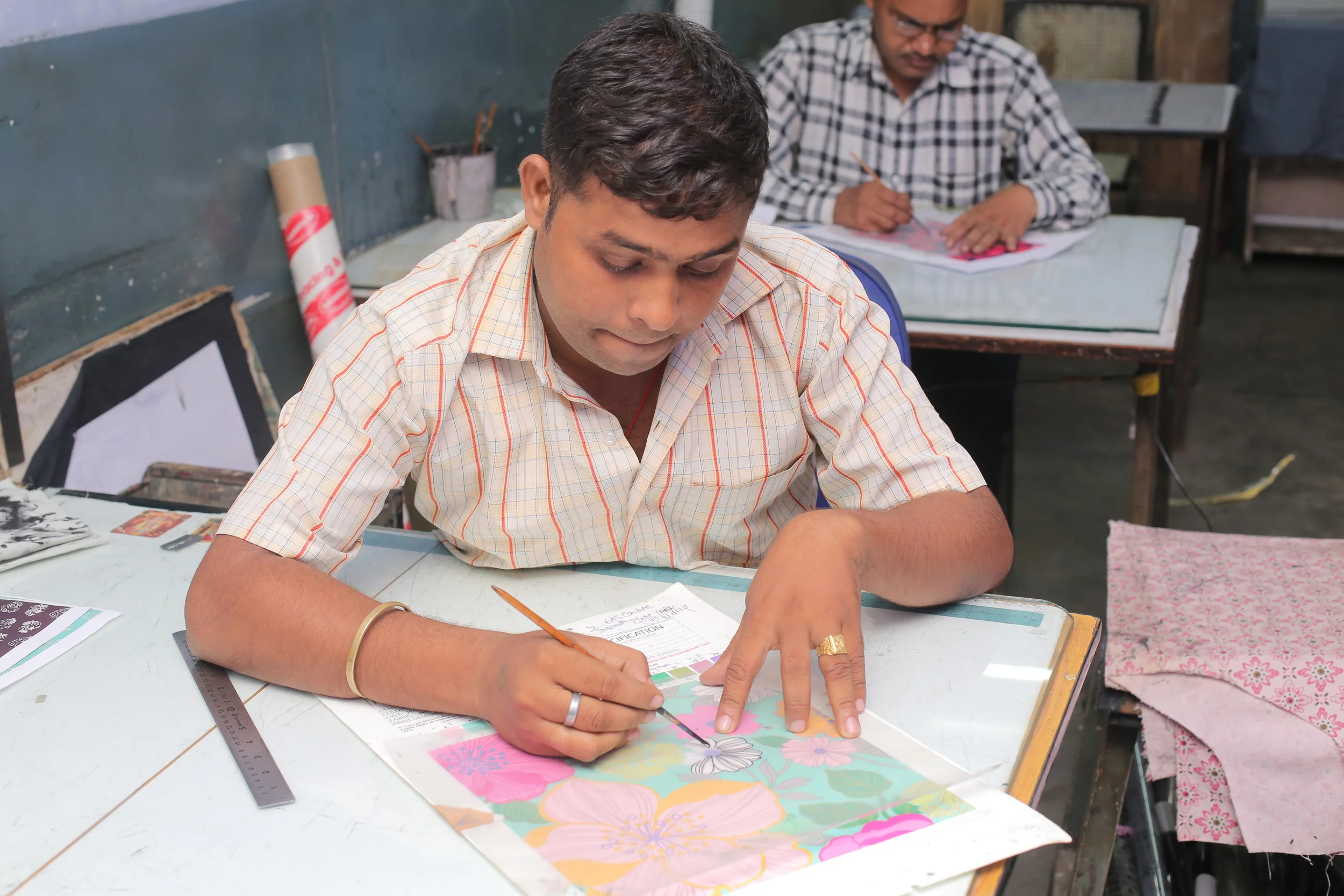 Man working on fabric printing with floral design in workshop, another man in background also working.