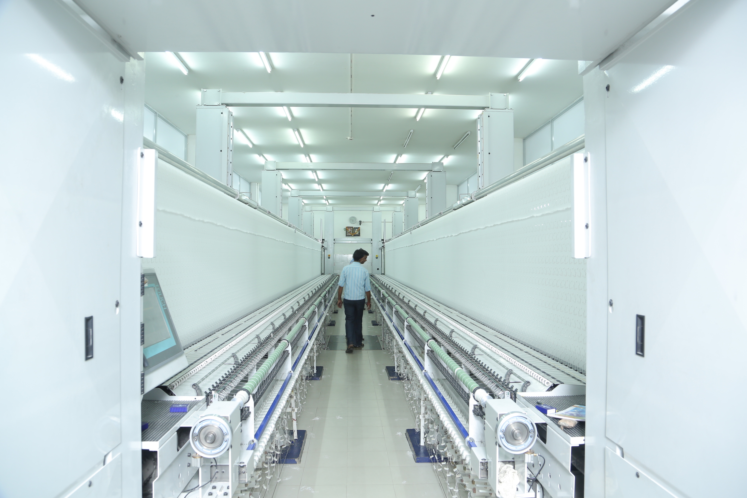 A person walks through a large, bright textile manufacturing or printing room with long, white machines on either side and a colorful painting on the back wall.