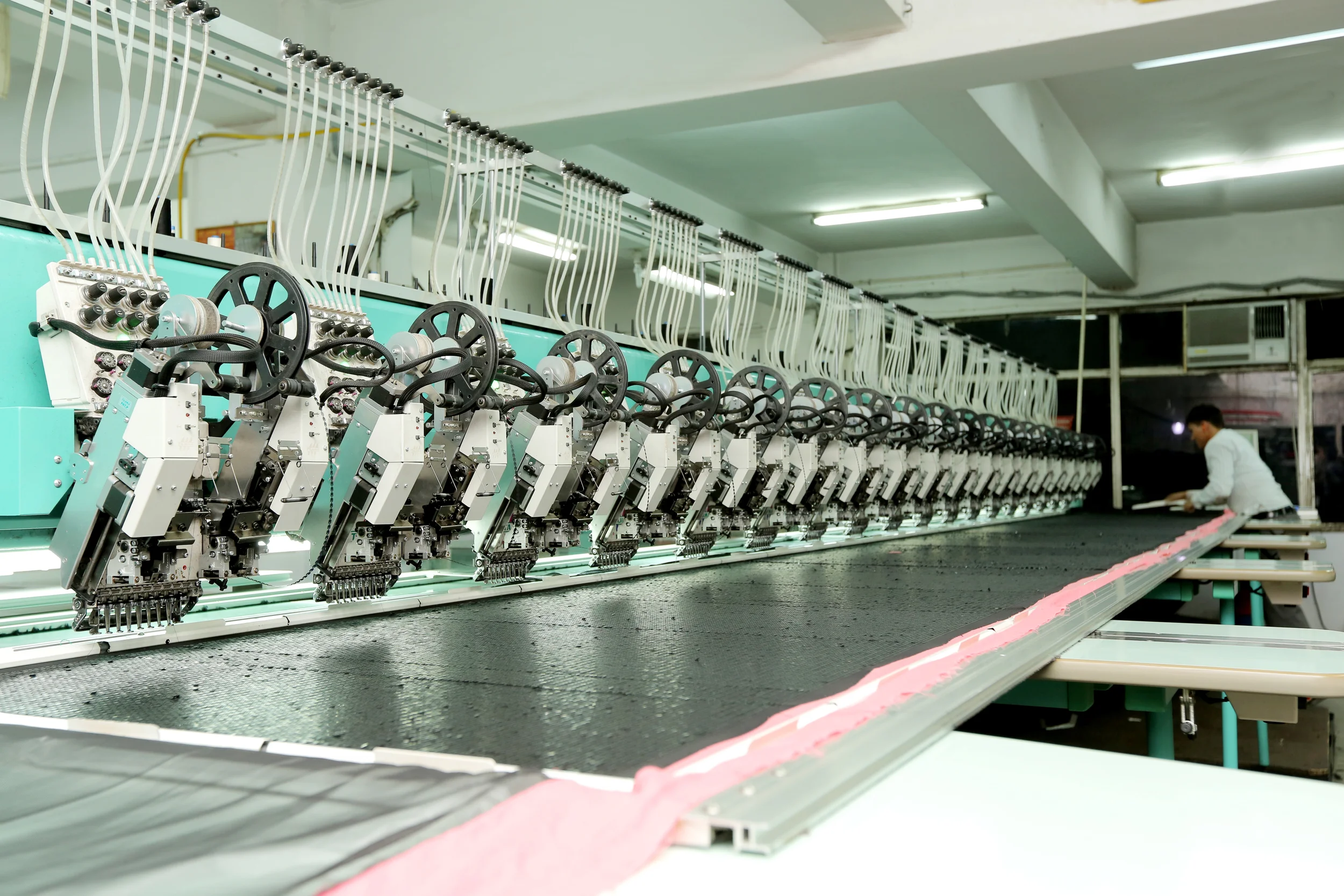 Industrial textile manufacturing machine with multiple embroidery heads and a worker inspecting pink fabric on a long table.