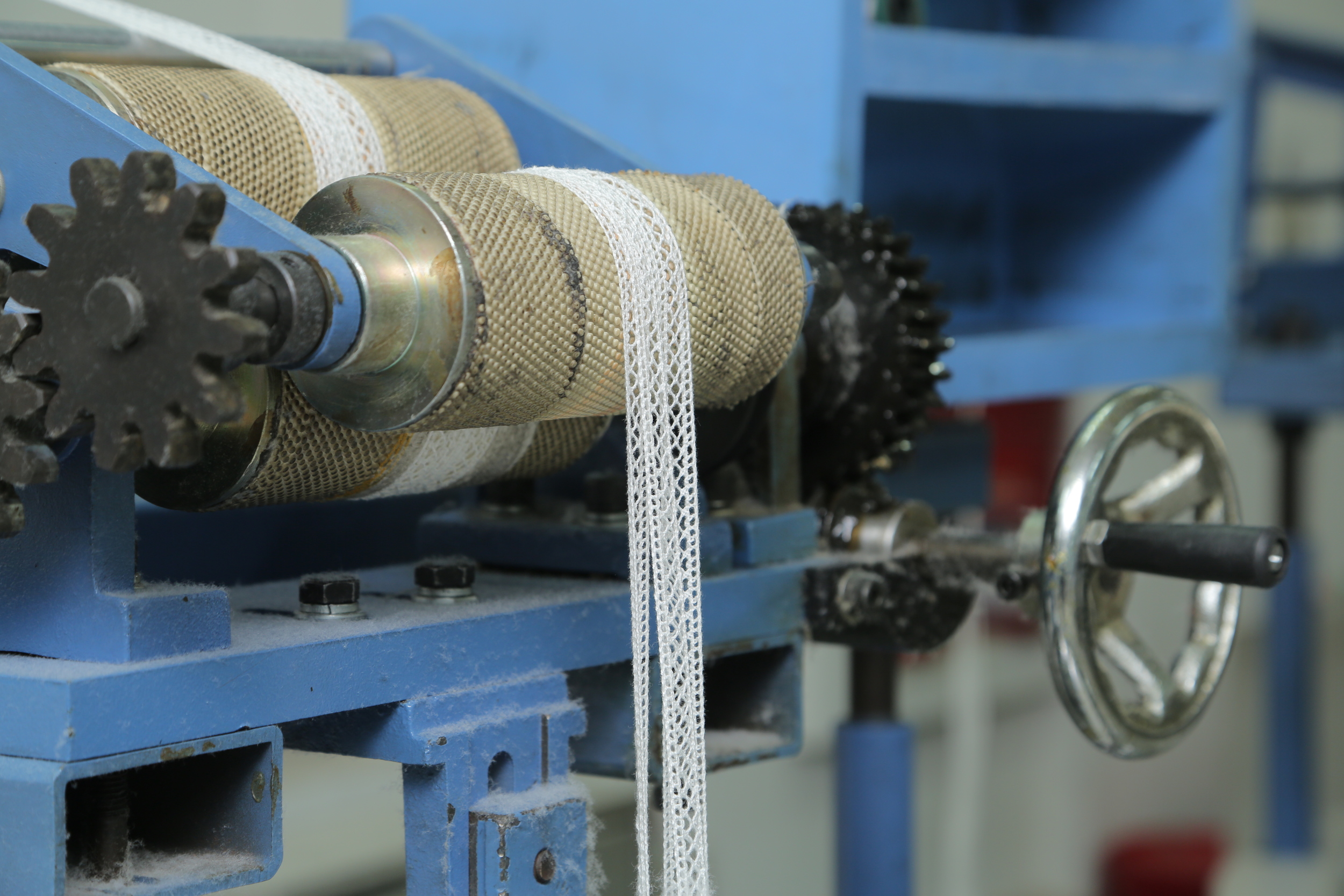 A machine with rollers covered in mesh fabric producing lace trim, with gears and a wheel to control the machine.