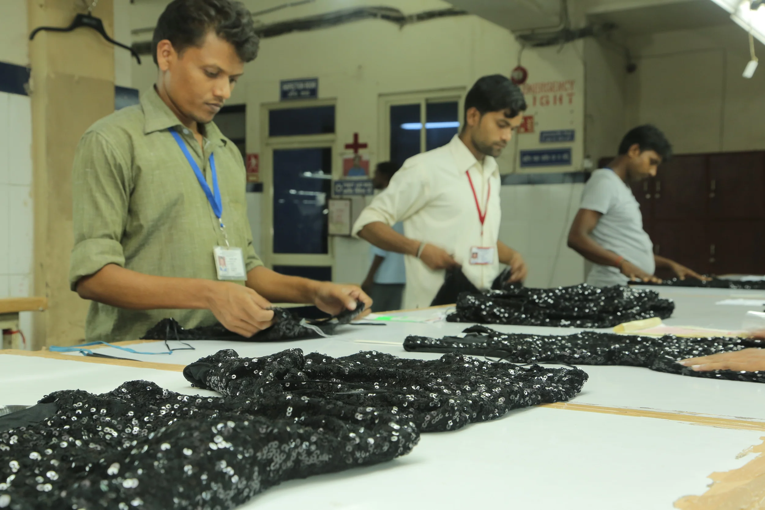 Three workers in white, beige, and gray shirts working at a table handling black sequined fabric in an industrial setting.