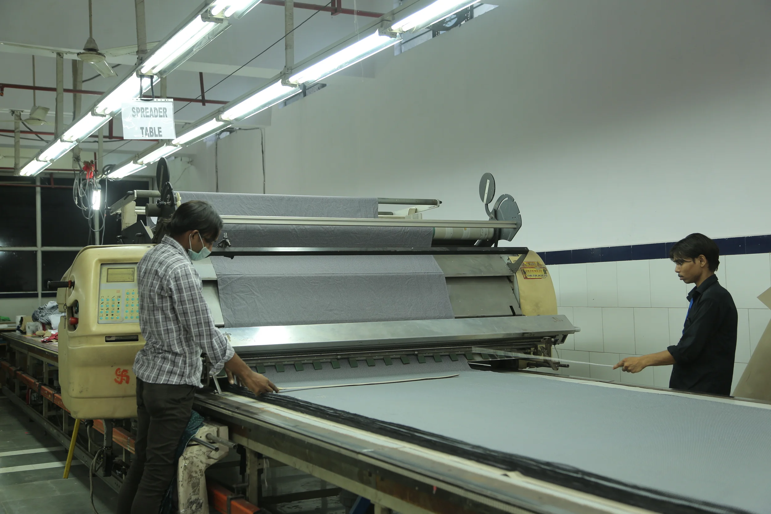 Two workers operating a textile machine in a factory, with one wearing a face mask and the other holding a tool, under bright fluorescent lights.