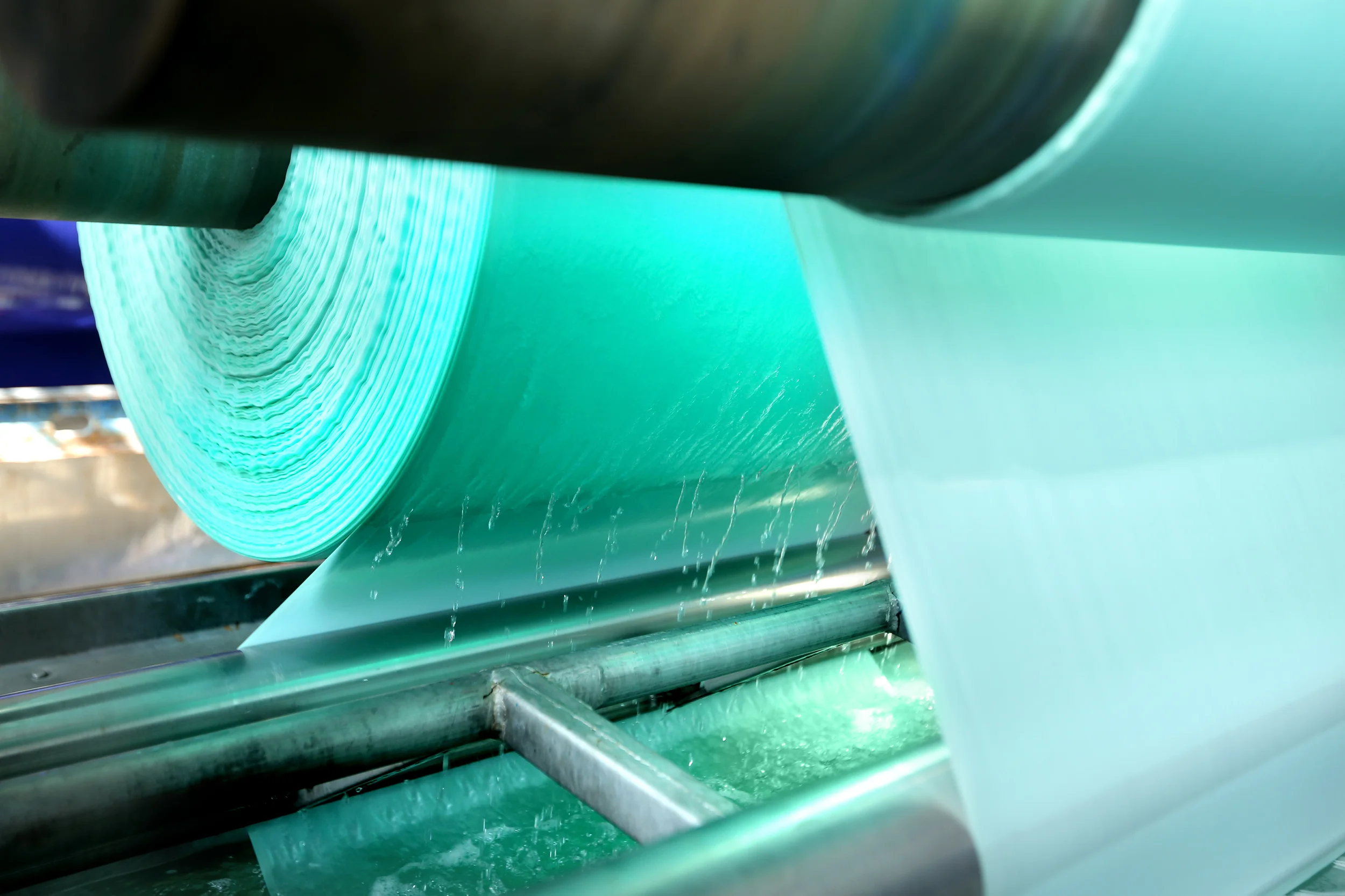 Close-up of large green industrial roll of plastic or film being fed through rollers with water droplet splashes.
