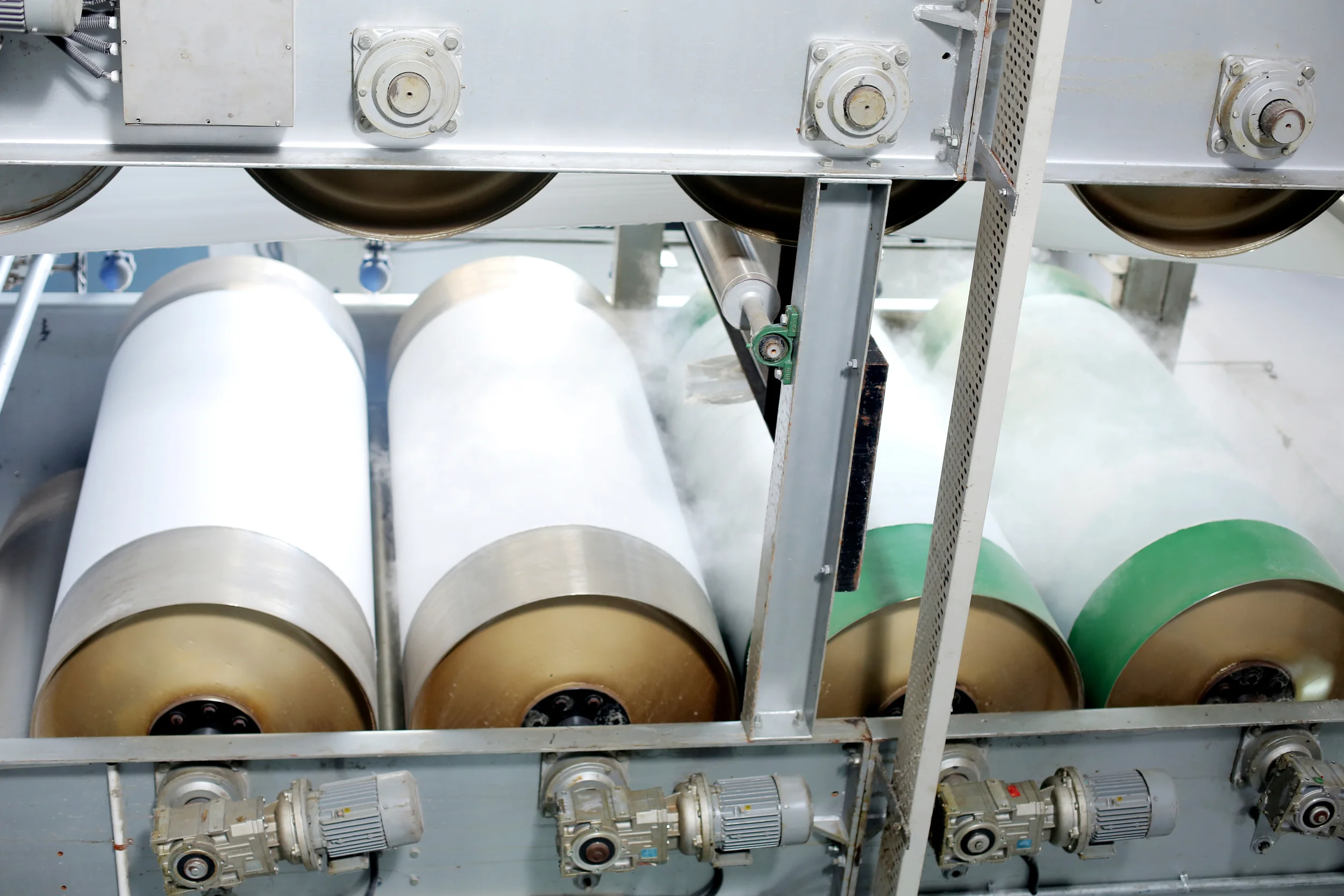 Industrial machine with large rollers, some with green or white covers, producing vapor or steam during operation.