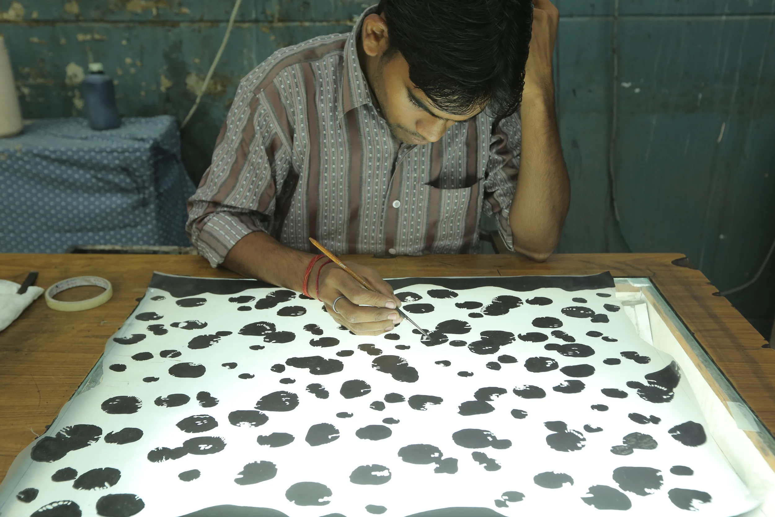 A man sitting at a wooden table, painting black ink blobs on a white paper illuminated from below, with a focused posture.