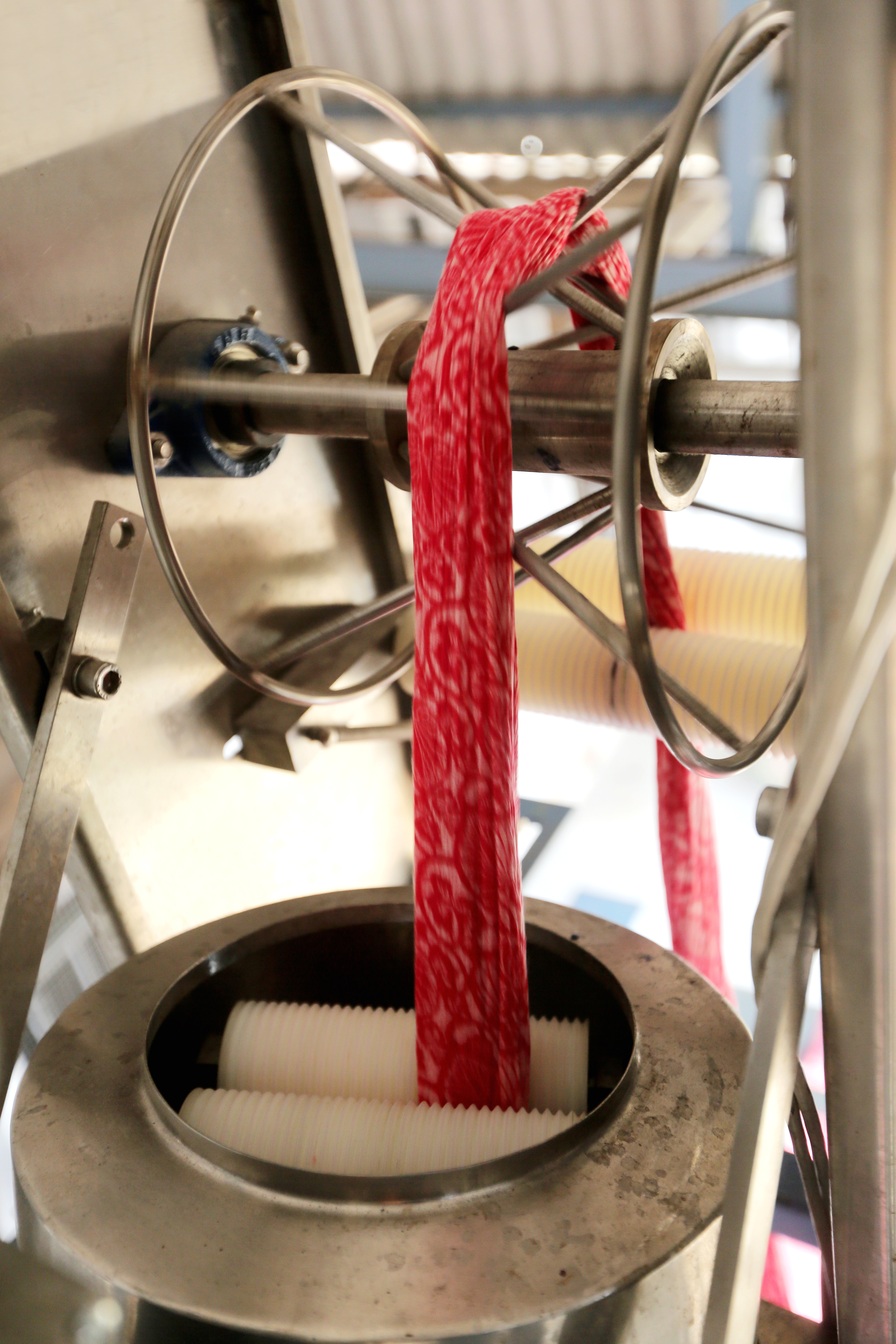 A close-up of a pasta-making machine extruding thin, flat pasta. A red strip of pasta is emerging from the machine's opening, with a yellow striped sheet lying beside it.