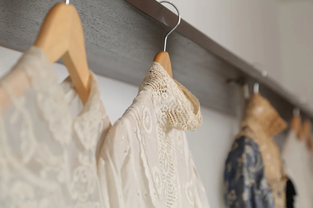 A row of lace and embroidered dresses hanging on wooden hangers on a metal rod in a closet.