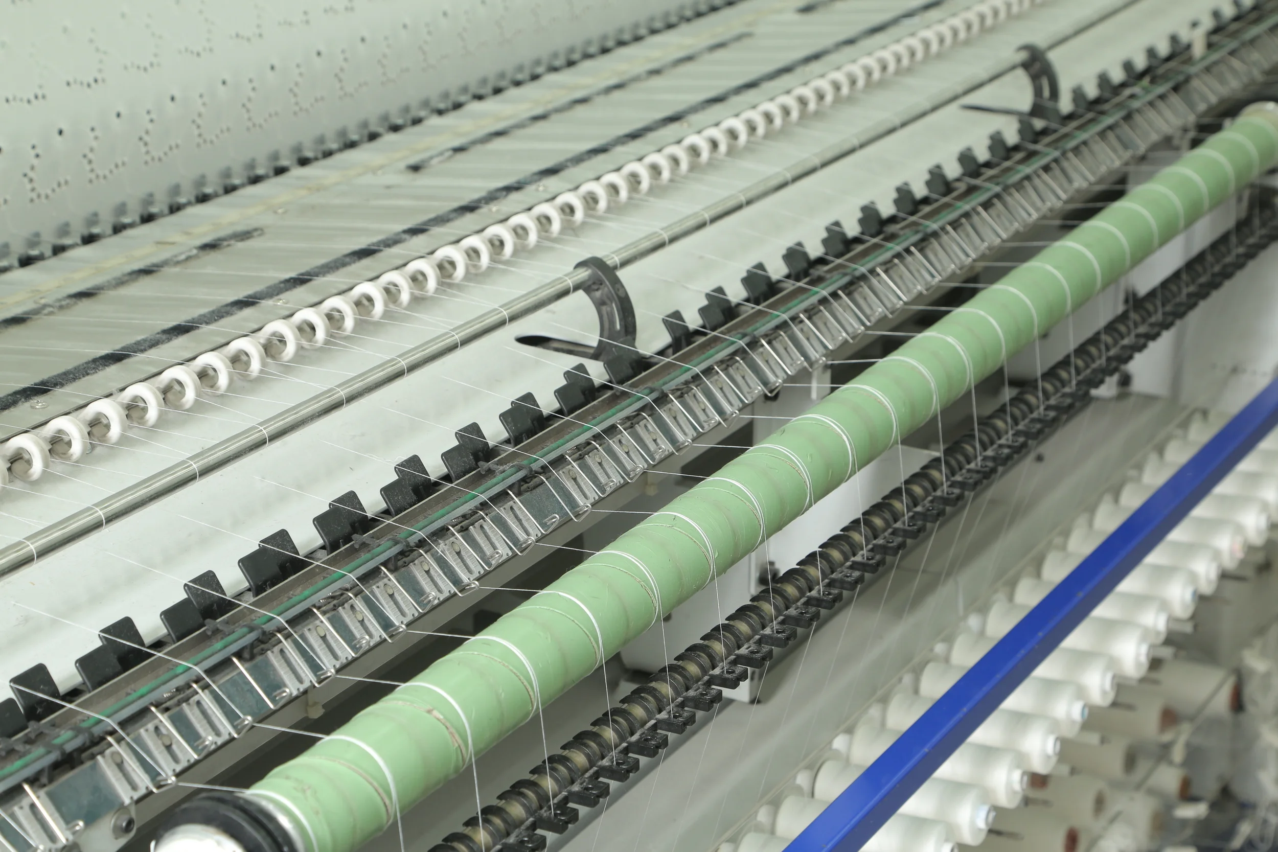 Close-up of a knitting machine with multiple rows of white, black, and green yarns being woven through various metal and plastic components.