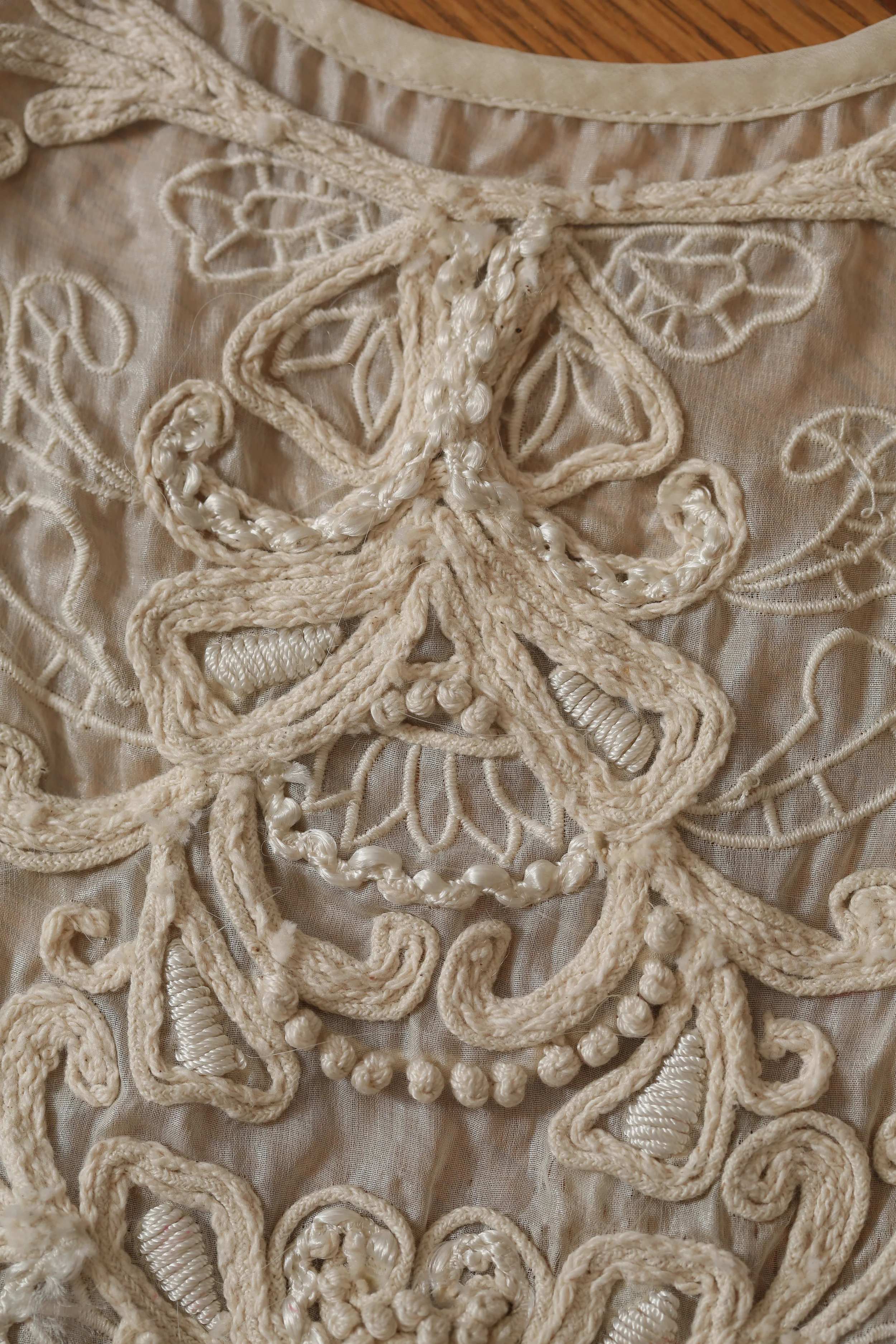 Close-up of beige embroidered fabric with intricate lace and corded embroidery patterns.