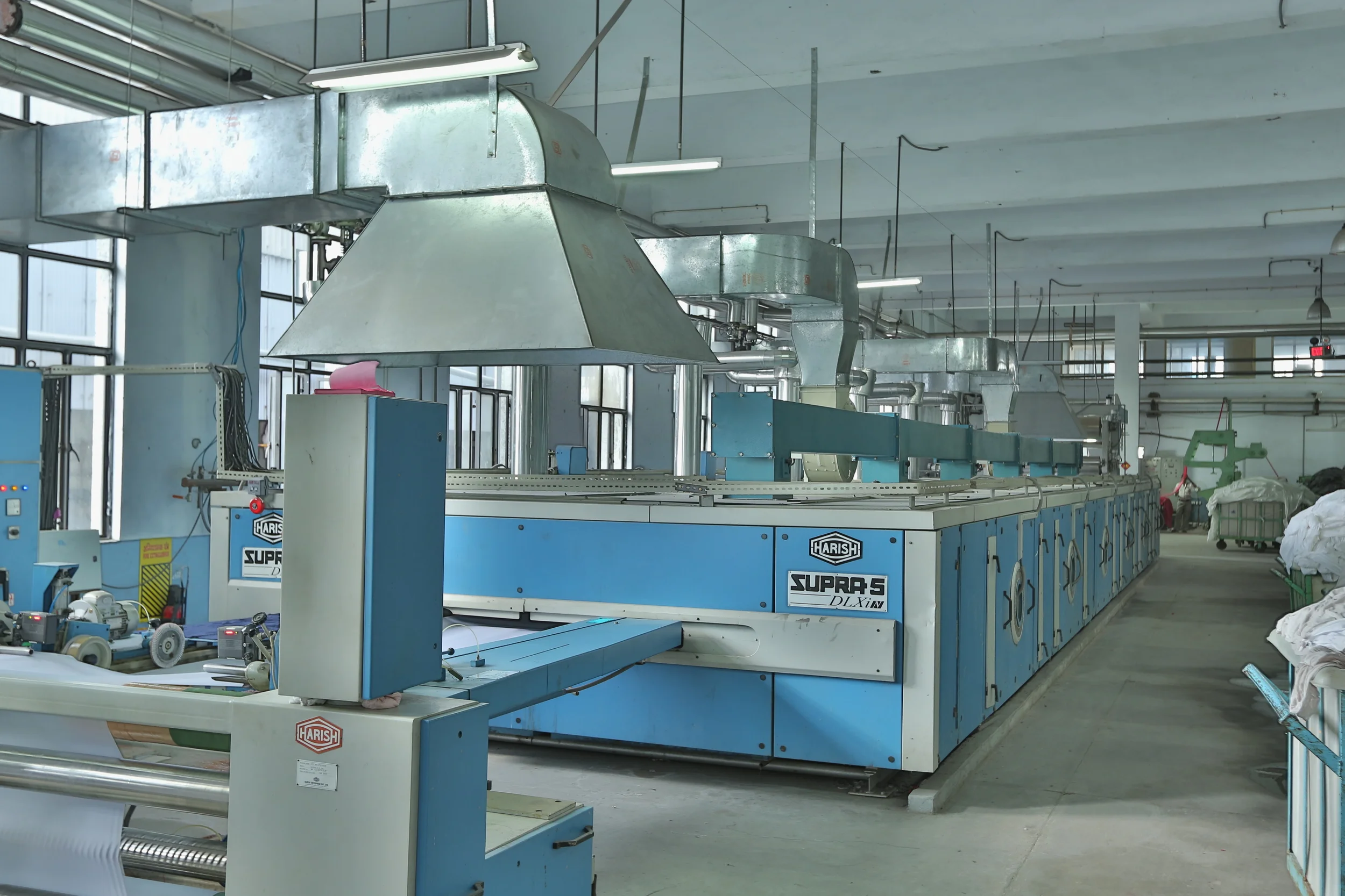 Industrial textile manufacturing machine labeled 'Harish Supra 5 DLX IN' inside a factory with pipes and ventilation system overhead.