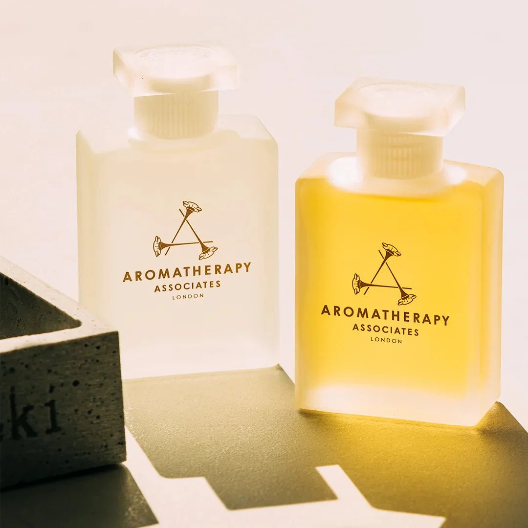 Two bottles of aromatherapy oils, one clear and one yellow, with black labels featuring a logo of two sticks with shoes, and the text "AROMATHERAPY ASSOCIATES LONDON."