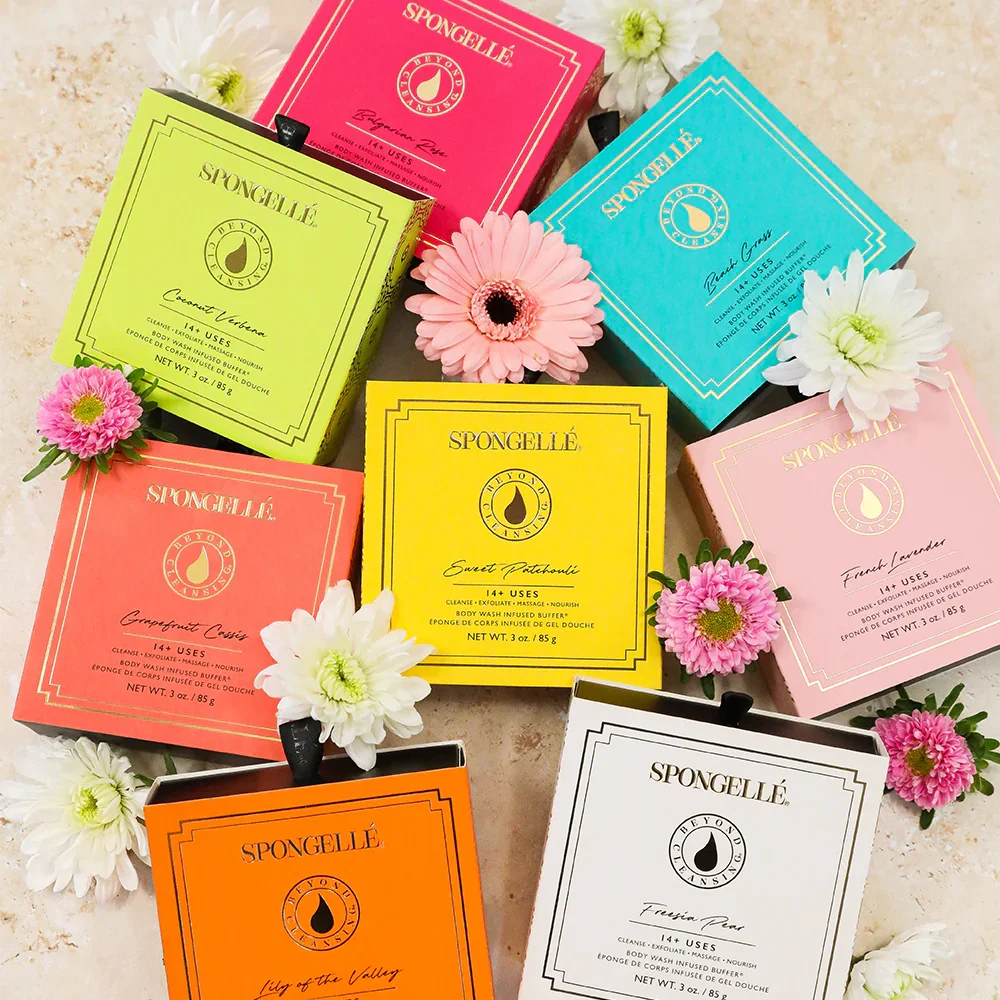Colorful boxes of Spongellé bath products arranged on a beige surface, decorated with pink and white flowers.