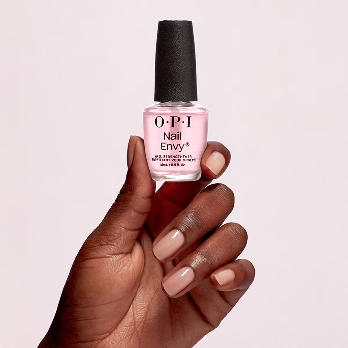 Close-up of a person holding a bottle of pink nail polish labeled 'O.P.I Nail Envy' with a black cap against a plain light background.