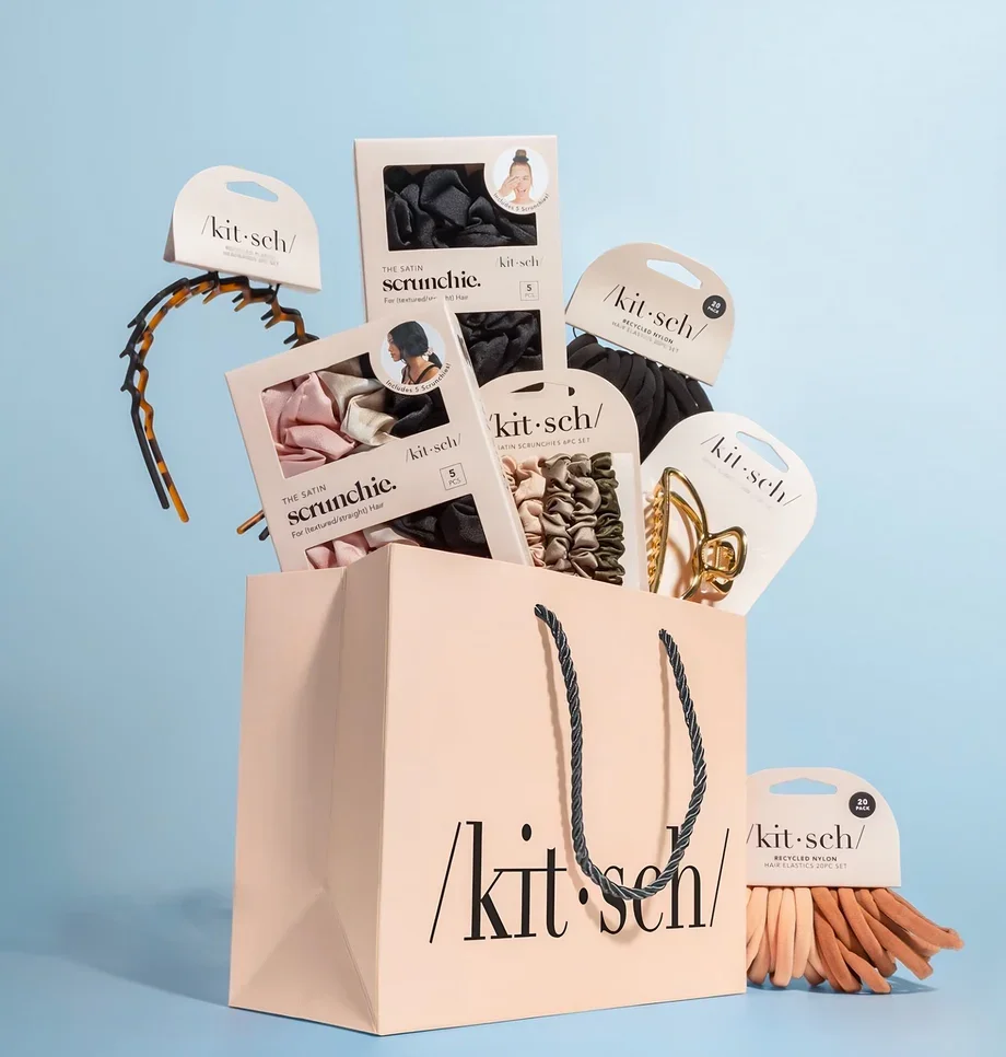 A collection of hair accessories, including scrunchies and hairbands, in a pink shopping bag with the brand name 'kit sch' on it. The accessories are in packaging and are arranged against a light blue background.