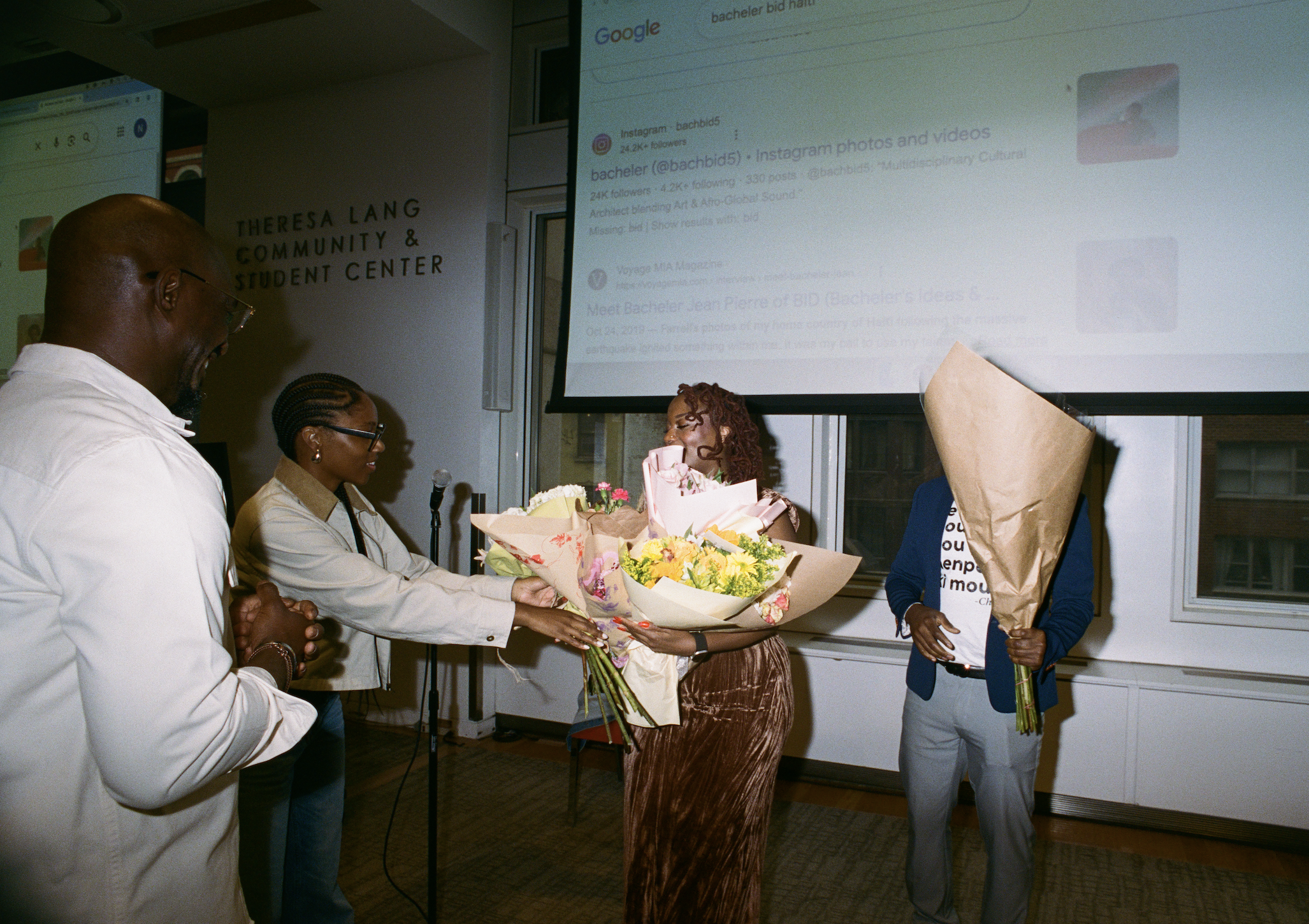 A woman receiving a bouquet of flowers during a celebration, surrounded by people at an indoor event in front of a screen displaying social media profiles and a sign that reads 'Theresa Lang Community & Student Center.'