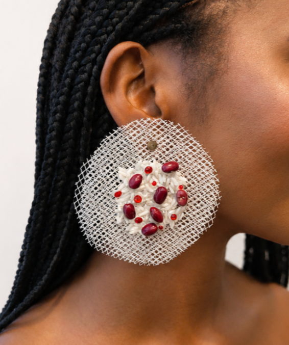 Close-up of a woman wearing a large, eating cupcake-shaped earring with white frosting and red and purple beads, against a plain background.