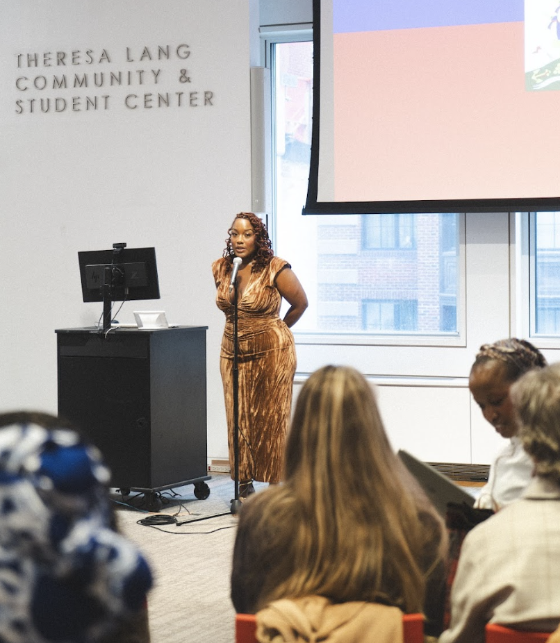 A woman in a bronze-colored dress speaking at a microphone during a presentation at the Theresa Lang Community & Student Center, with an audience seated and a large screen behind her.