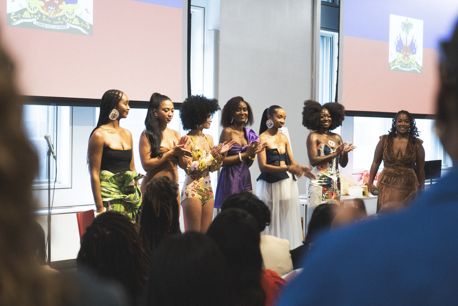 A group of women on stage in colorful outfits, clapping and celebrating in front of an audience, with two large screens displaying similar images in the background.