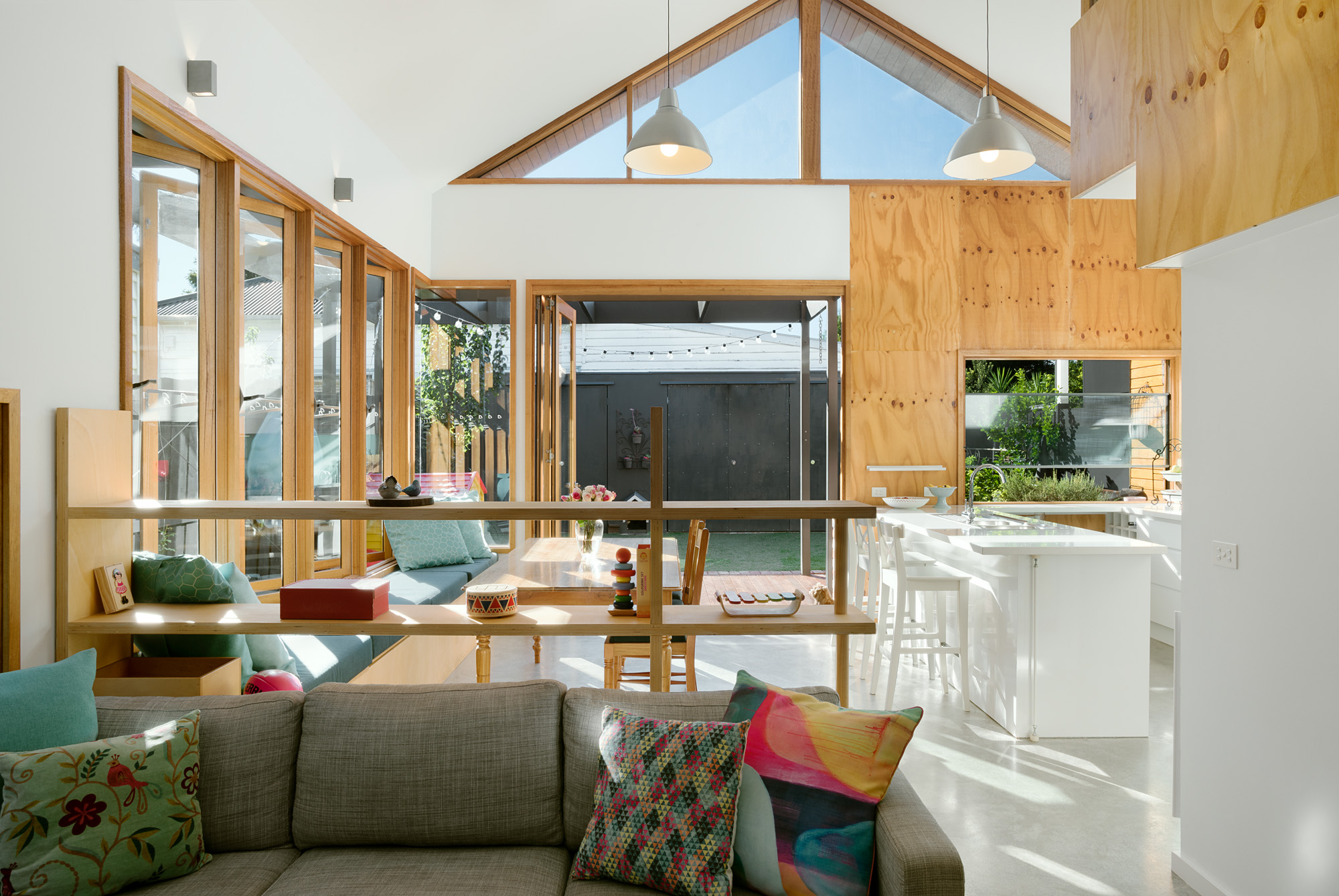 Open-plan living, dining and kitchen space beneath a raked gable window, with a plywood-lined mezzanine and built-in timber shelving