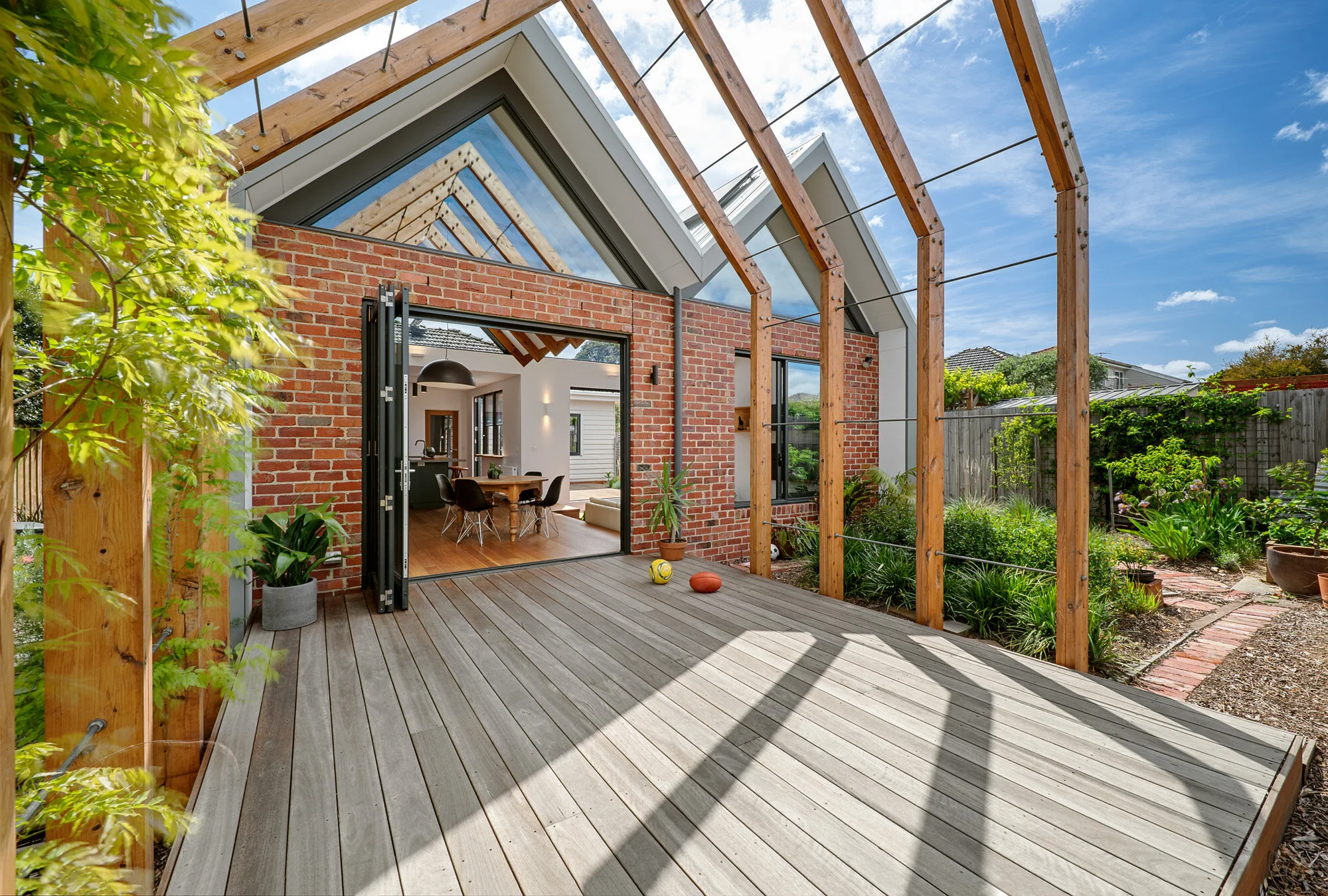 Timber deck beneath the vine-trellis pergola, looking into the dining room through open glazed doors in the brick facade