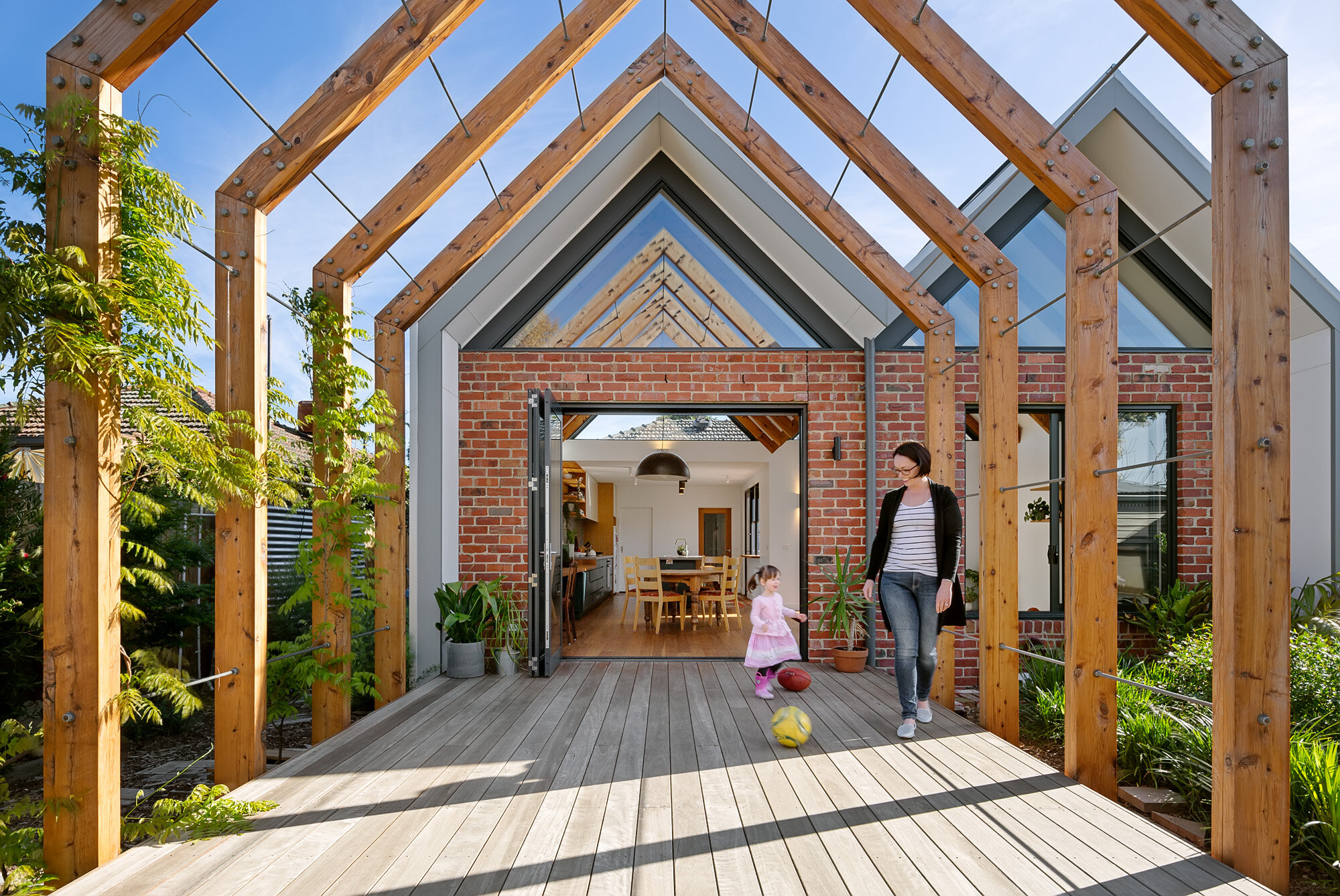 A parent and child at the open glazed doors beneath the timber pergola, framed by the recycled brick gable wall