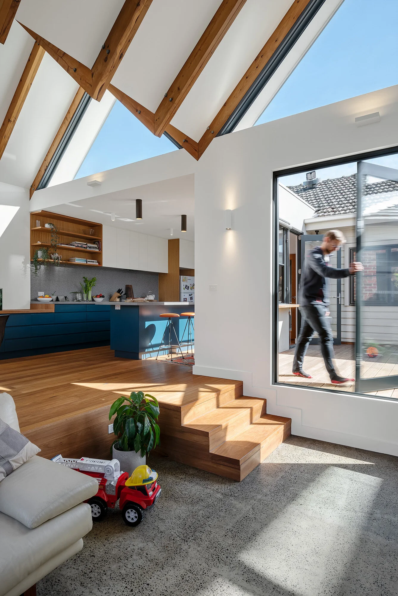 Split-level living space where a timber stair steps up from the sunken concrete-floor lounge to the raised timber-floor kitchen