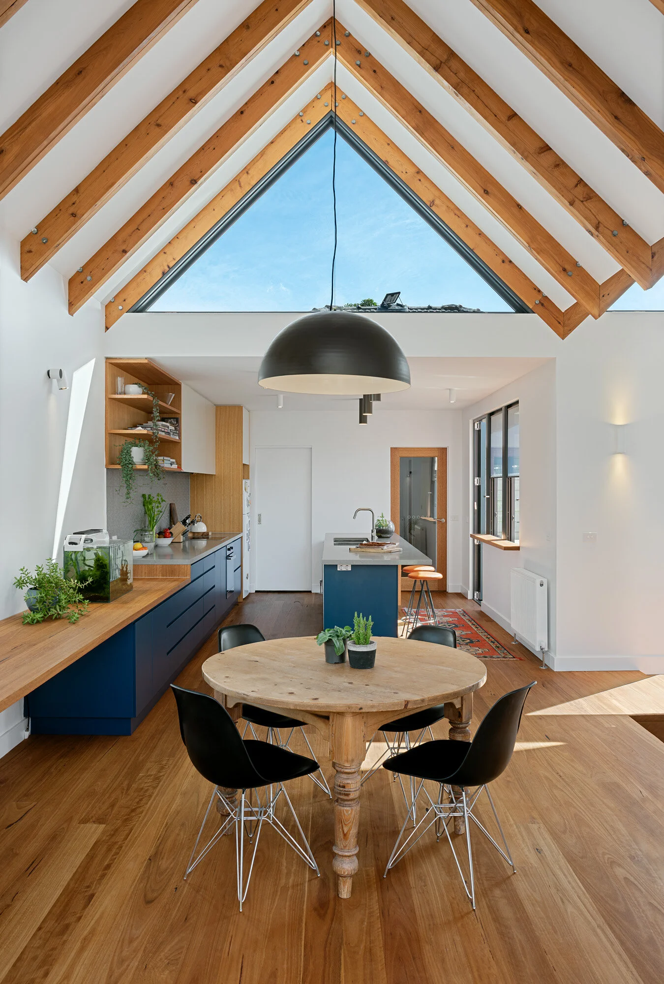 Dining table centred beneath a full-height triangular gable window, with the kitchen's exposed timber rafters above