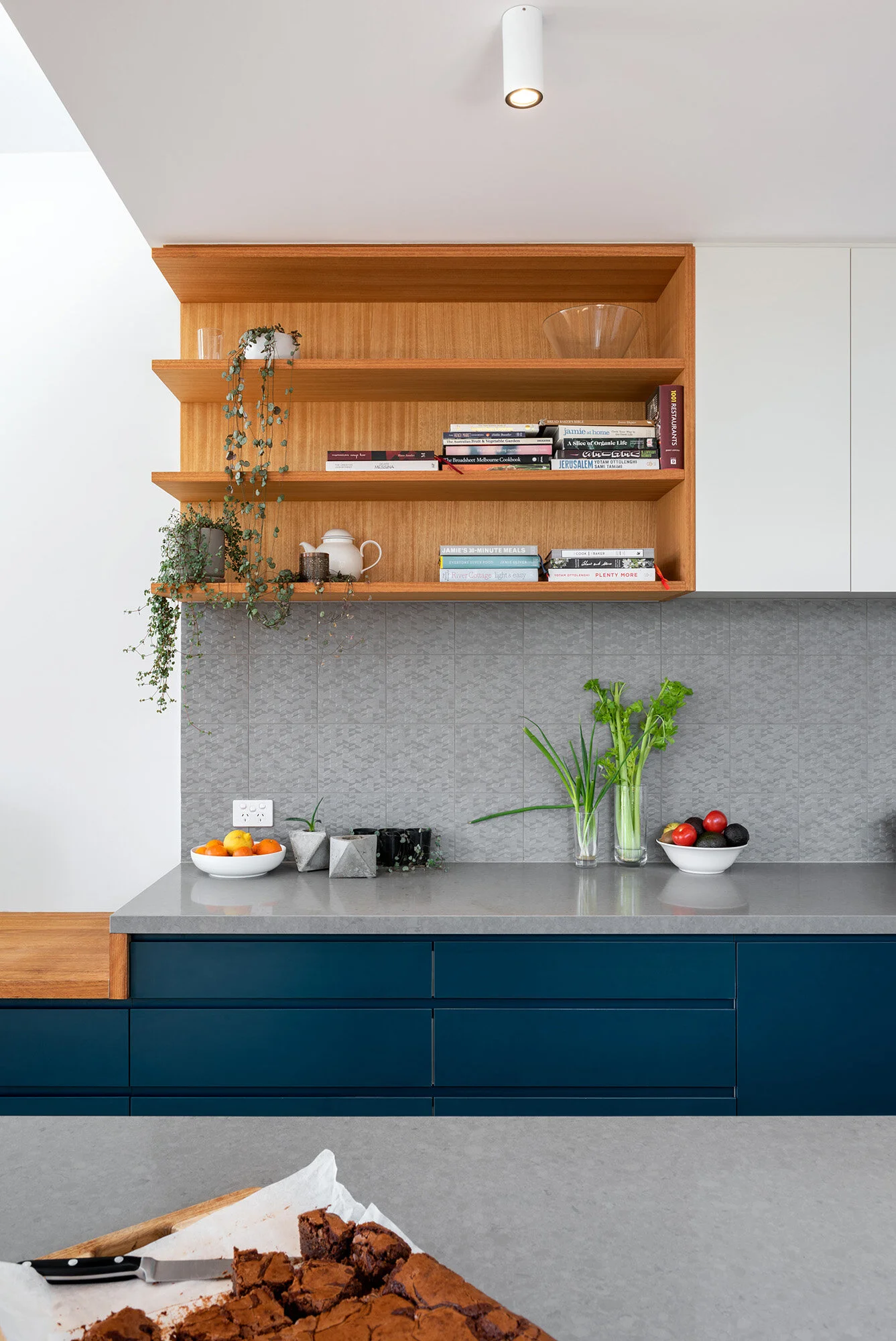 Kitchen shelving detail with cookbooks and trailing plants above a patterned tile splashback and navy joinery