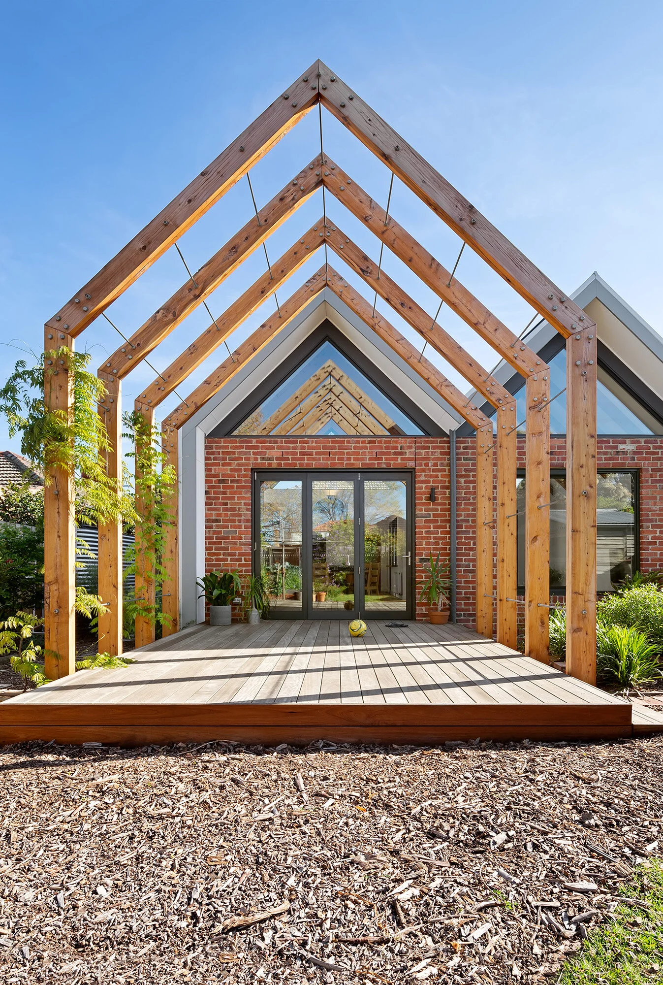 Repeated gable-form timber pergola trained with climbing vines for seasonal shading, leading to a recycled brick extension
