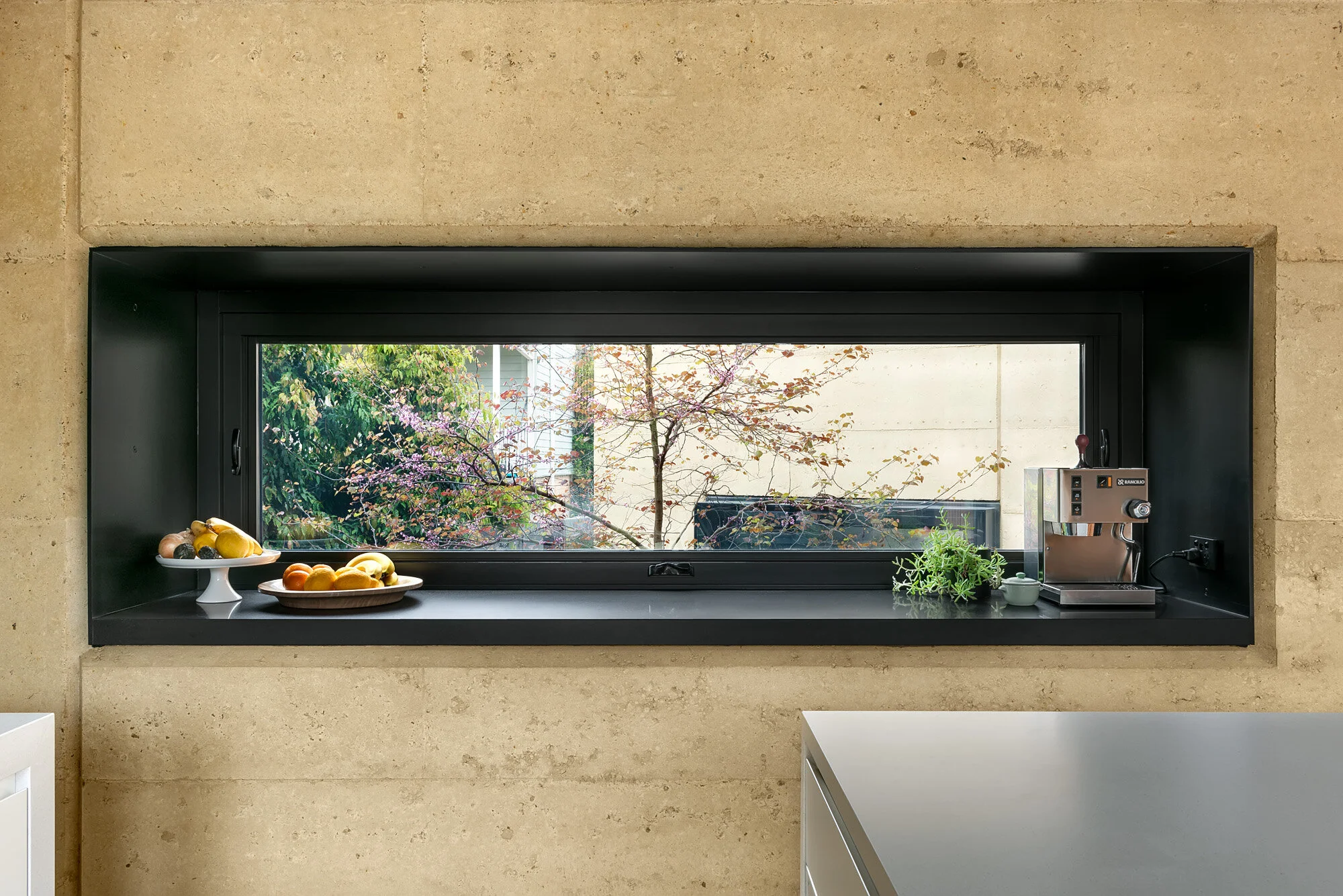 Deep kitchen window reveal framing view to courtyard through rammed earth wall.