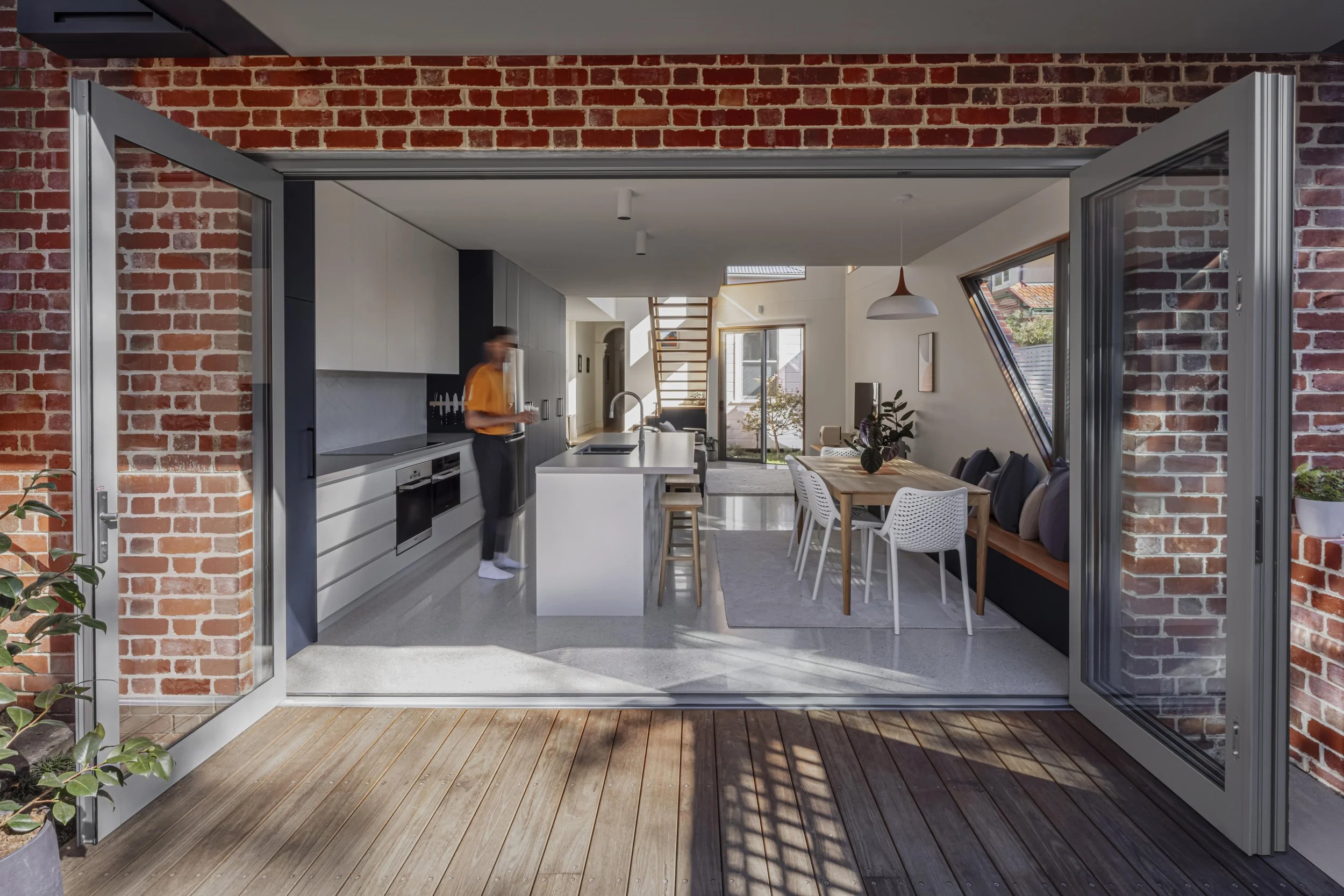 Glazed bifold doors in a recycled brick opening, connecting a white kitchen and dining space to a timber deck
