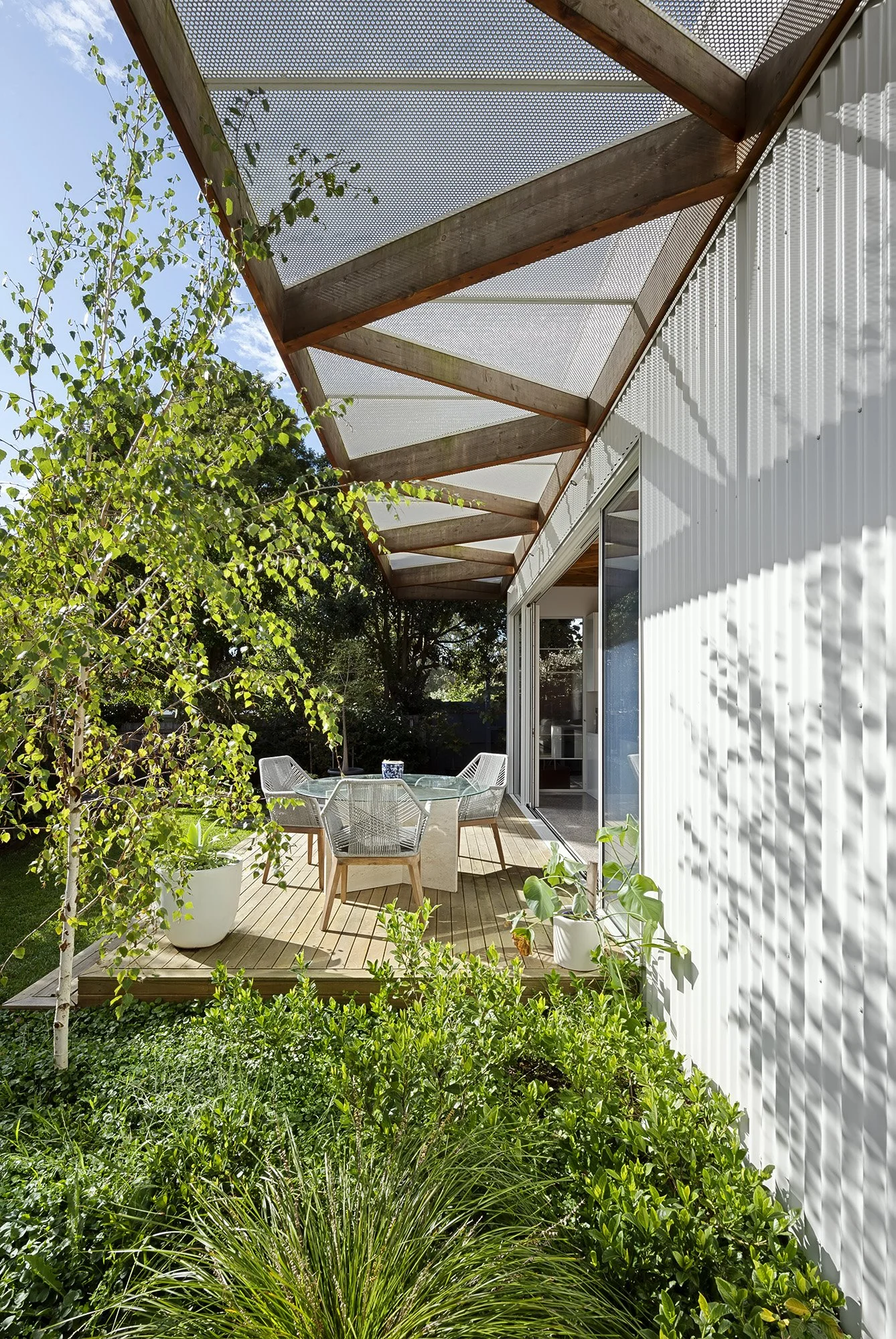 Exterior deck shaded by a perforated steel pergola on angled timber rafters, bordered by garden planting