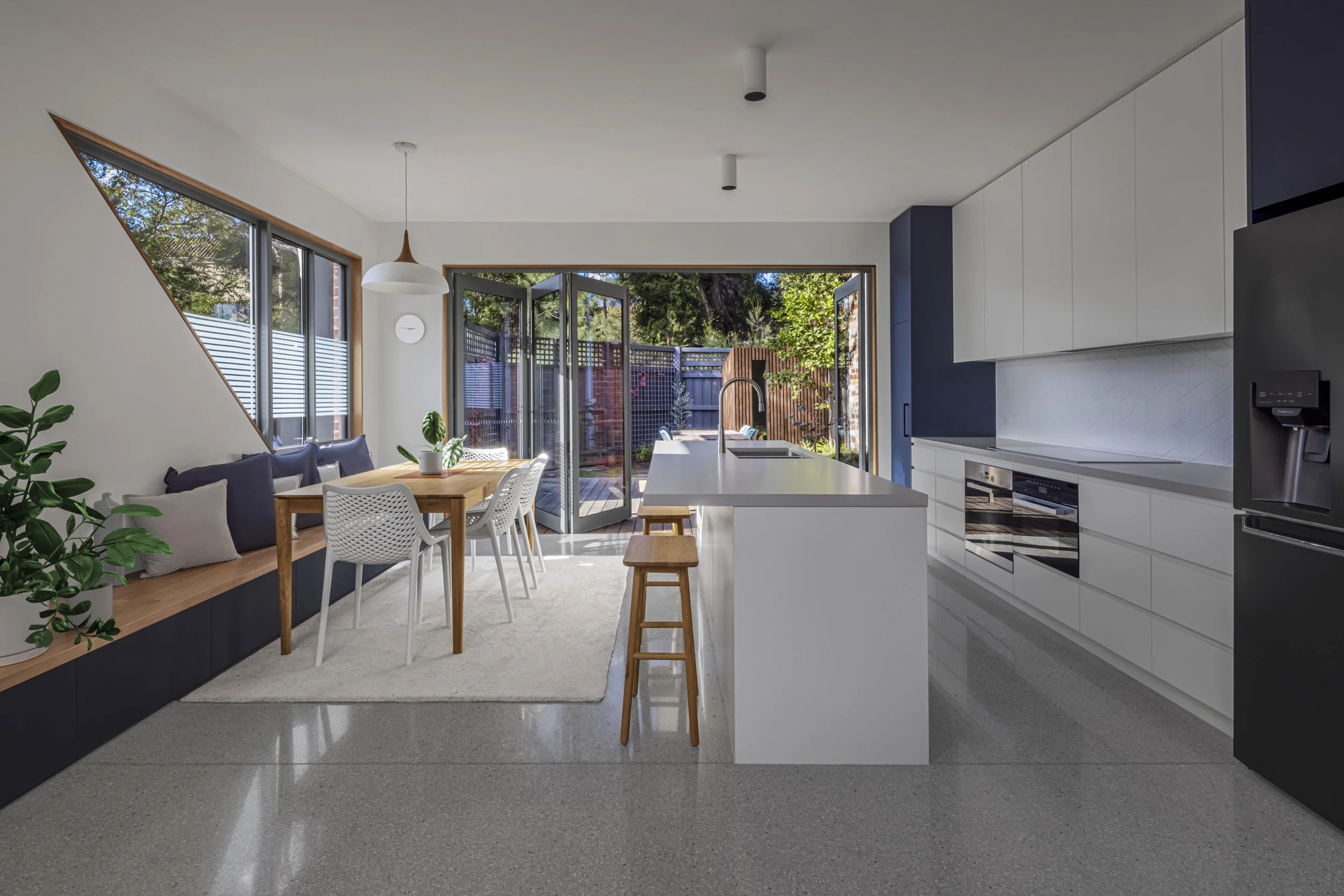 Open-plan kitchen and dining area with a built-in window seat beneath an angled skylight window, opening onto a timber deck and screened garden