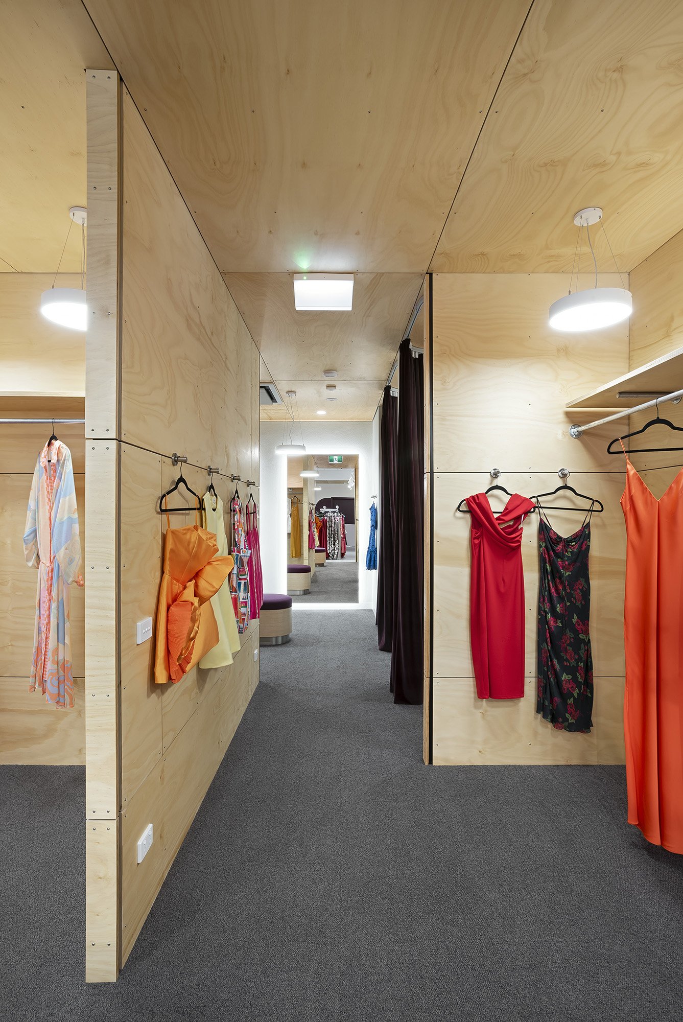 Plywood-lined fitting room corridor with mirrored end walls and dresses hung on hooks between curtained changing bays