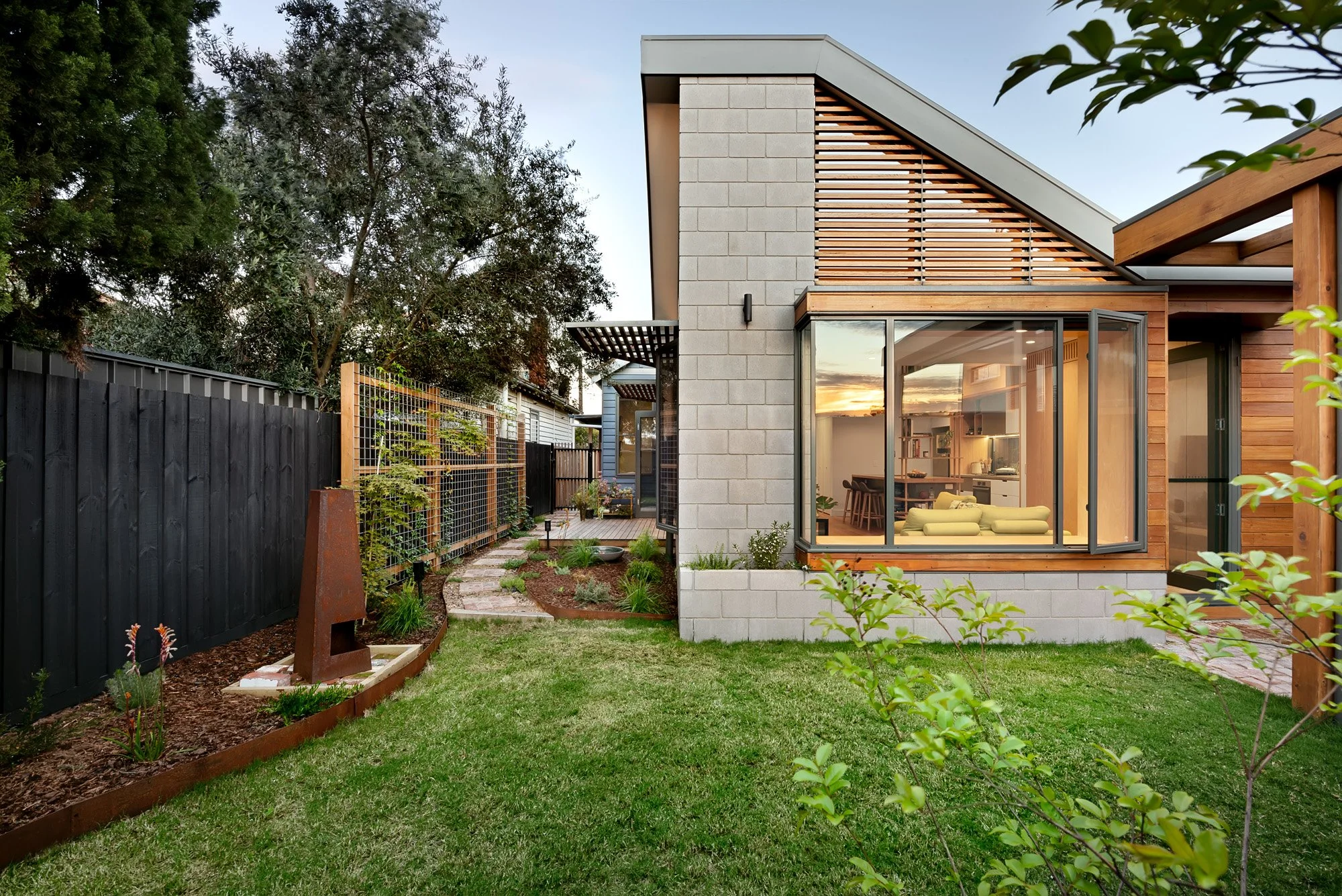 Wider dusk view of the raked-roof pavilion with timber battens and concrete block, bordered by a native garden path